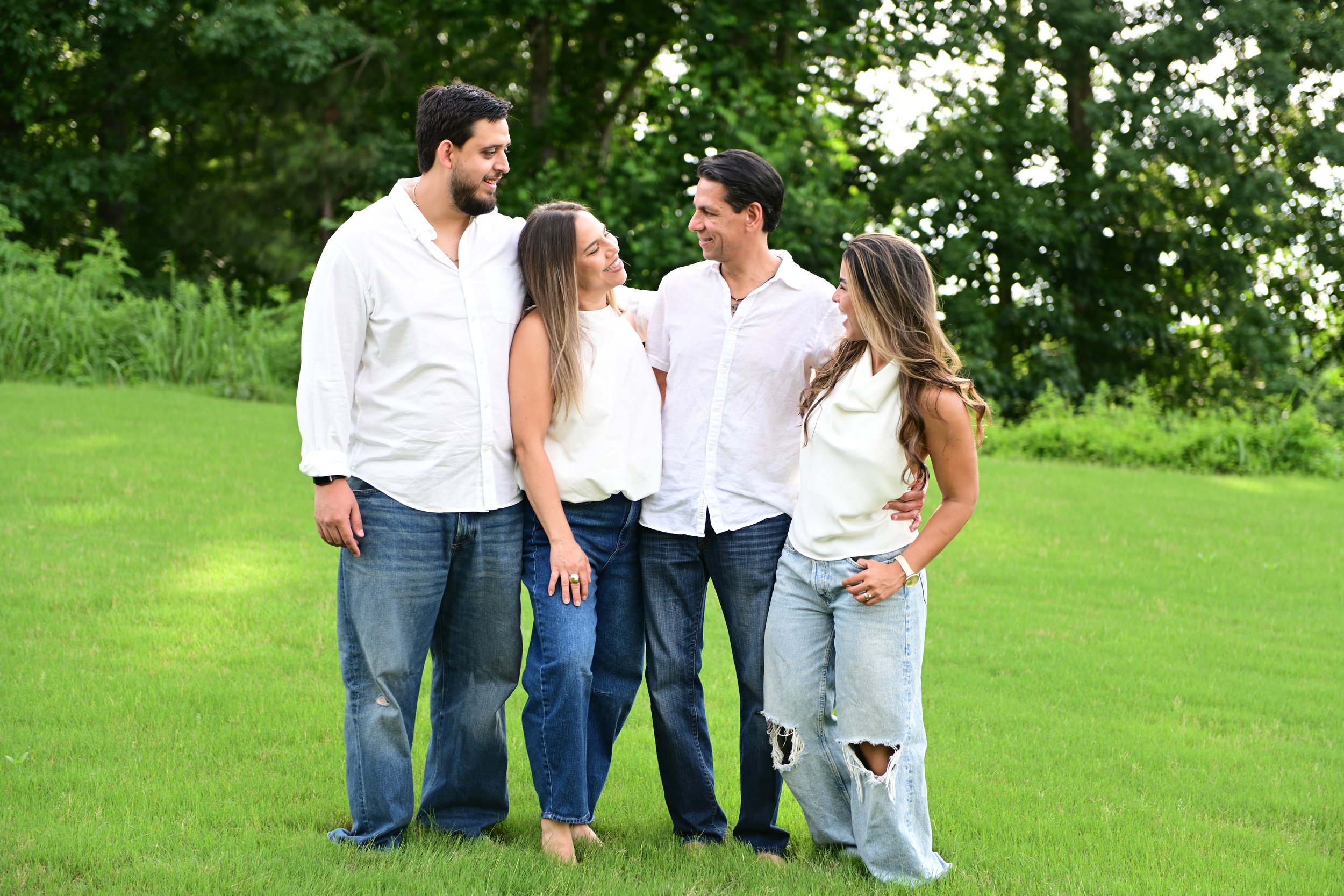 Group of five friends standing close together outdoors on a grassy area, smiling and engaging in conversation surrounded by trees.