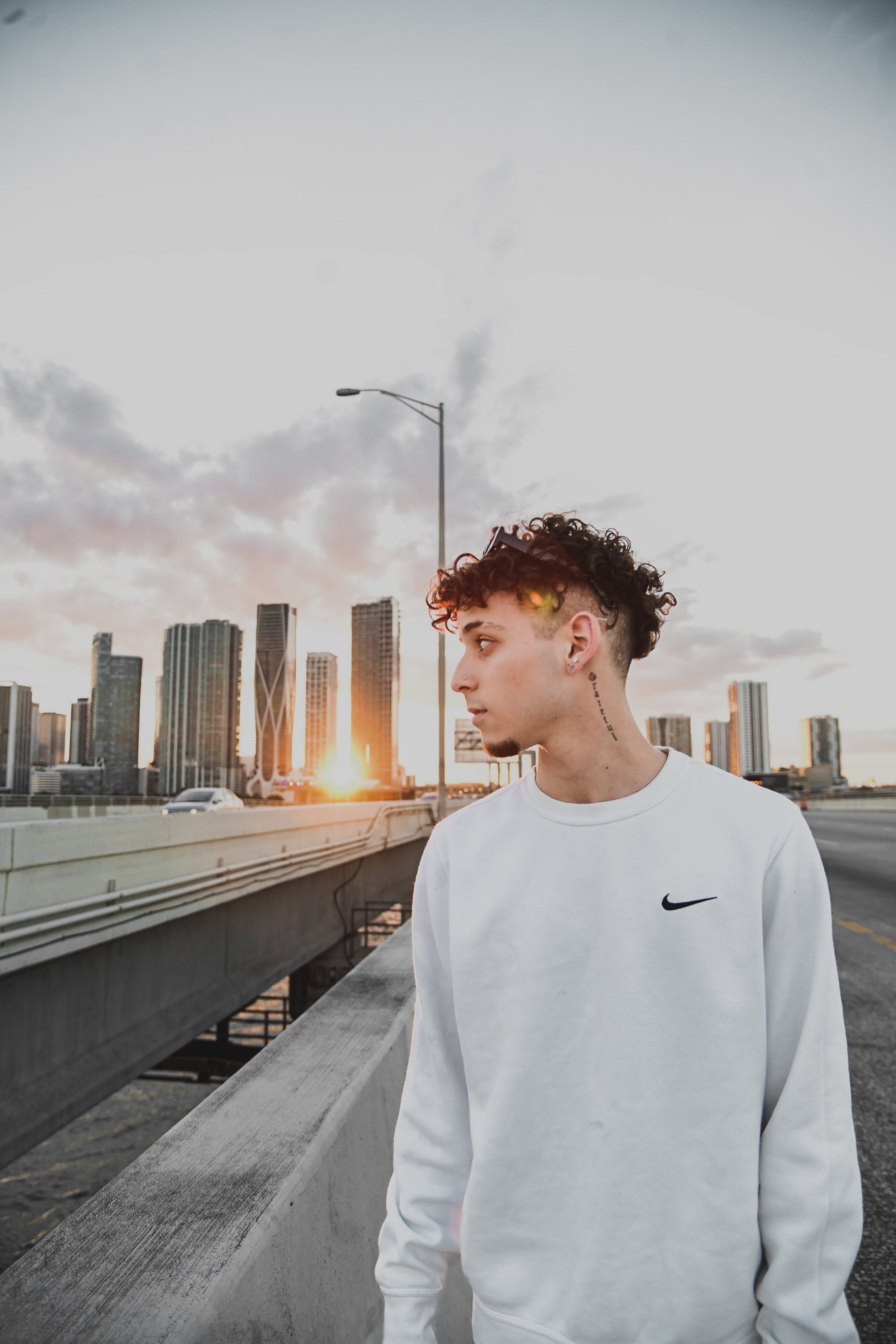 A young man with curly hair and earrings standing on a rooftop parking garage with a city skyline in the background during sunset, wearing a white Nike sweatshirt.