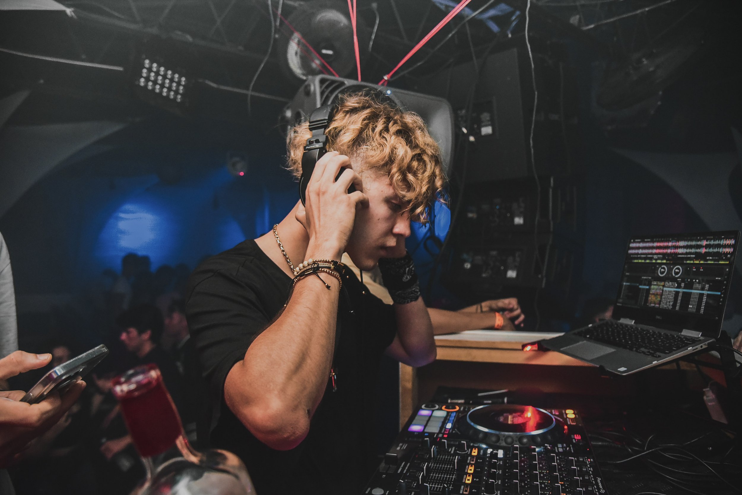 A DJ with curly hair wearing headphones and bracelets, standing at DJ equipment including a mixer and laptop in a dimly lit nightclub or event space with a crowd in the background.