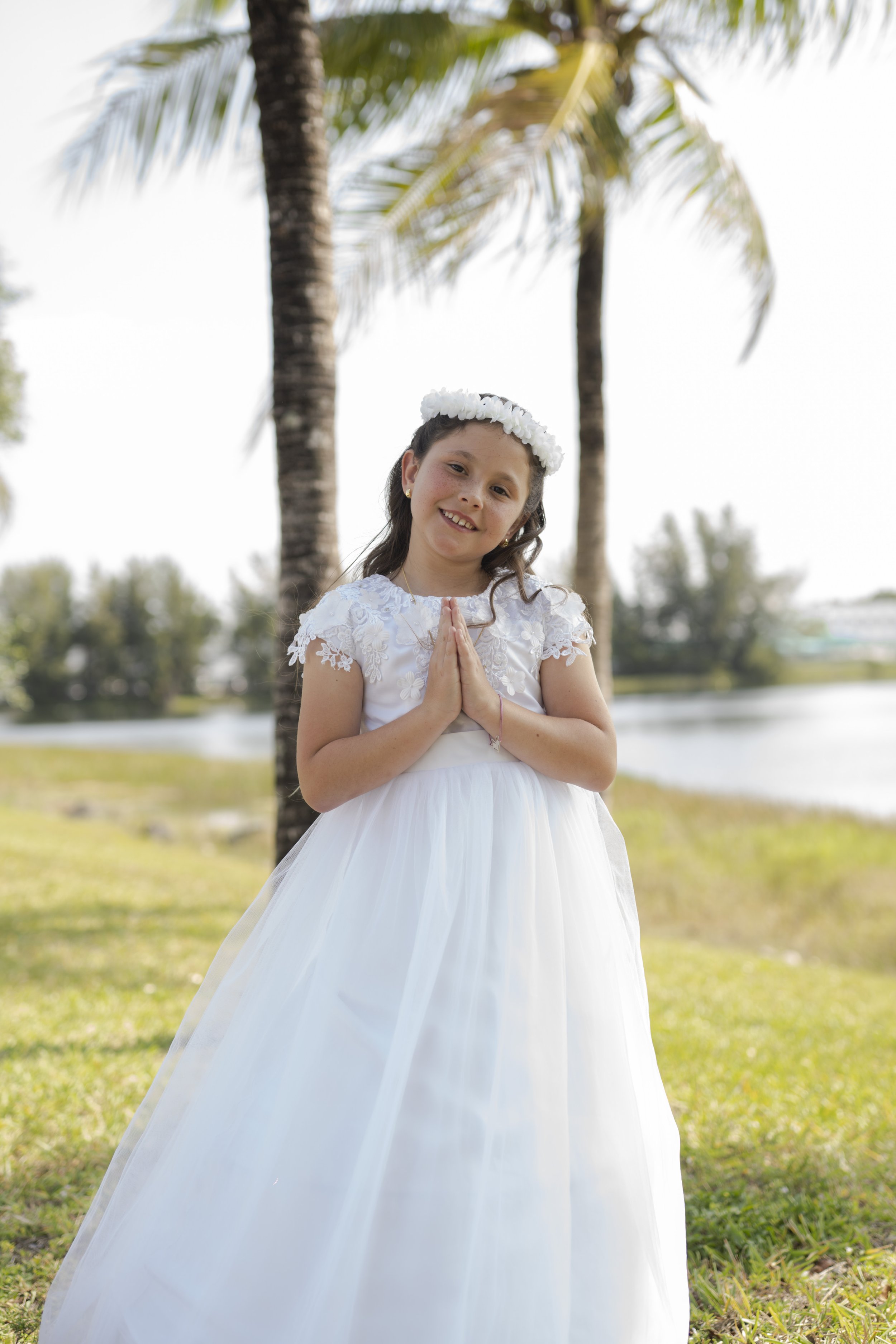 A young girl in a white dress with lace details, wearing a flower crown, standing outdoors with palm trees in the background. She is smiling and has her hands pressed together in front of her chest.