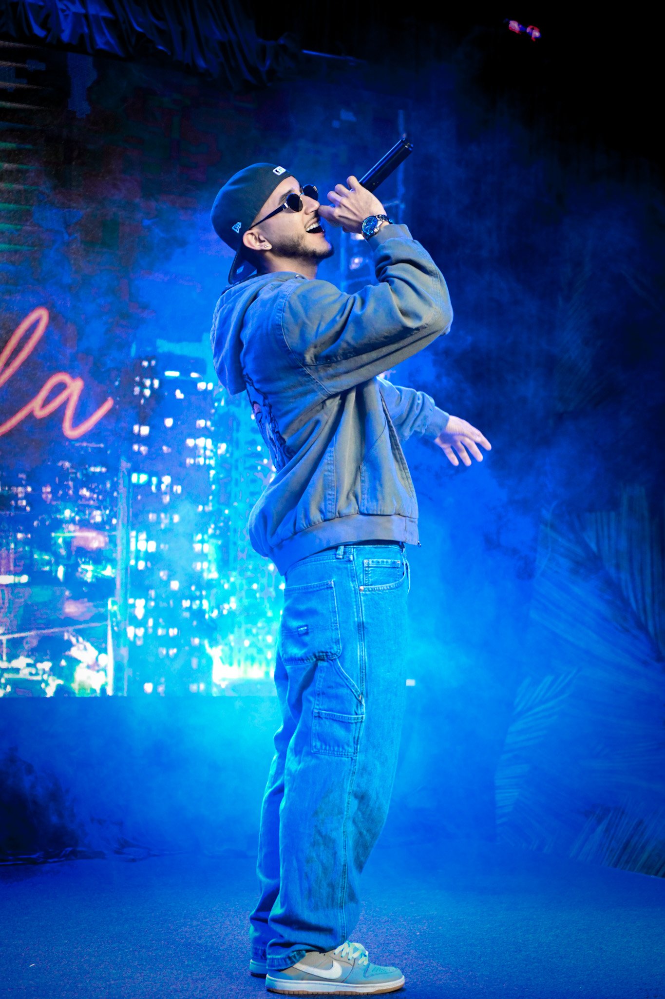 Young man wearing sunglasses, a black cap backwards, a gray hoodie, and jeans, singing or rapping into a microphone on stage with blue and purple lighting.