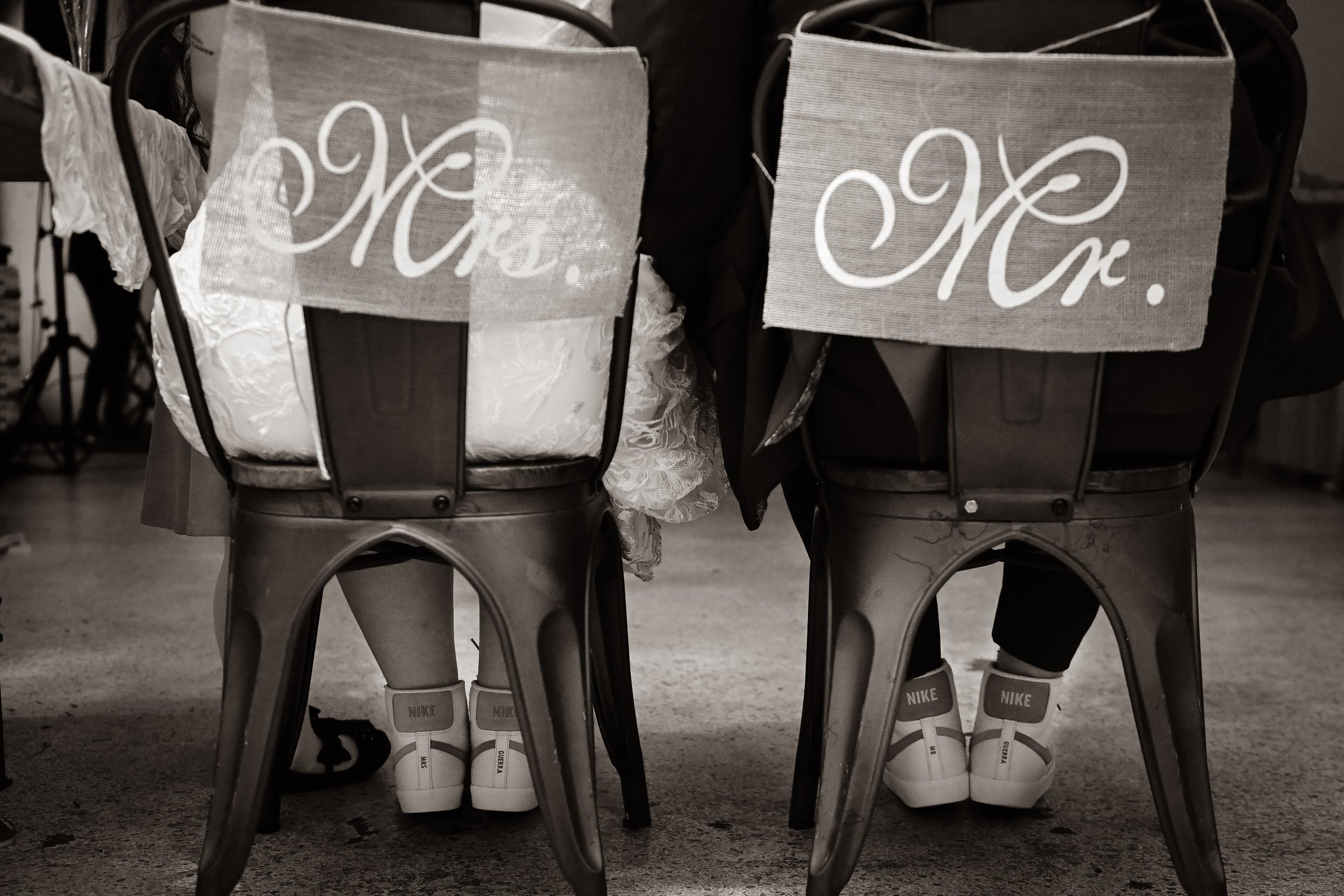 Two chairs with signs that say "Mr." and "Mrs." on the back, and people sitting on them wearing casual shoes, including Nike sneakers.