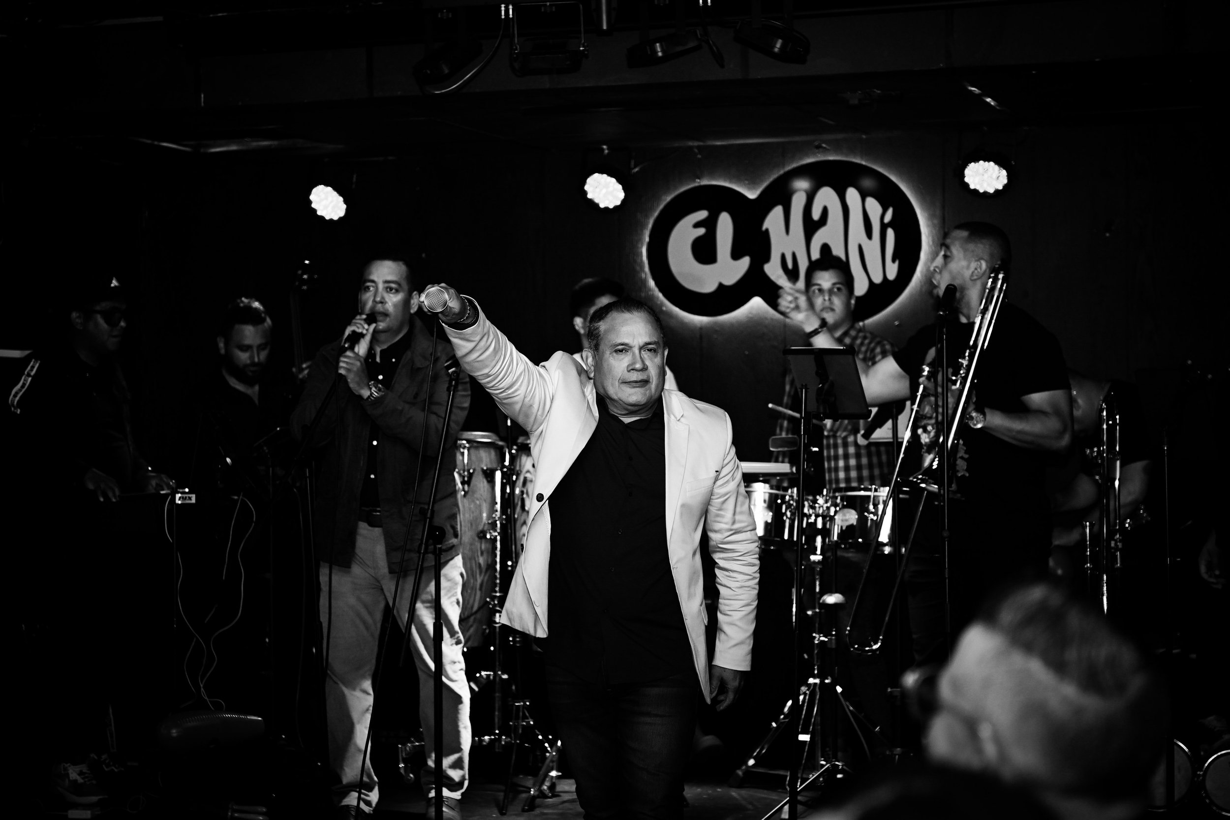 A band performing on stage at El Maní, with several musicians playing instruments and one man in a white jacket speaking or singing into a microphone.