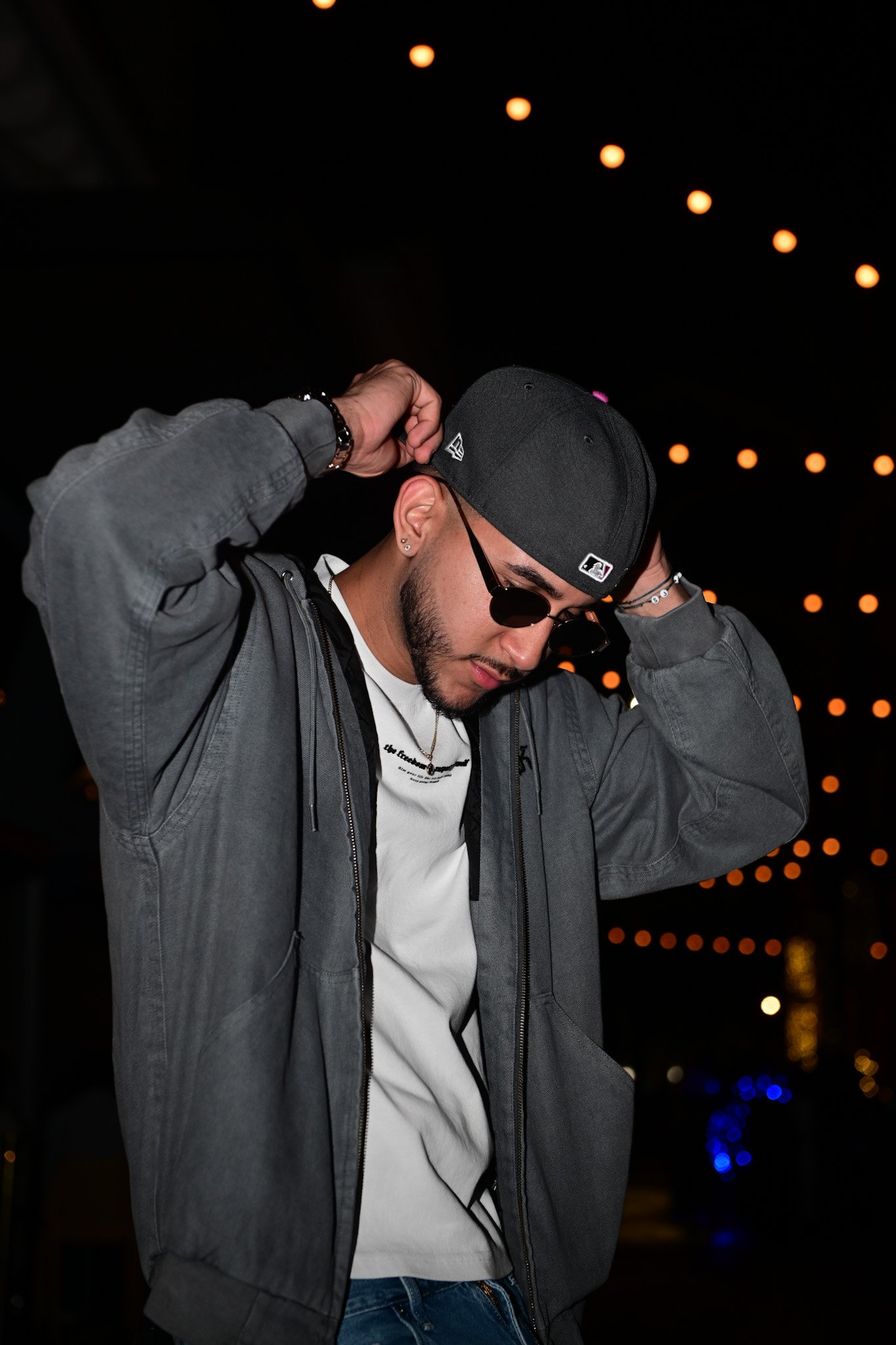 Young man wearing a grey jacket, white t-shirt, black baseball cap, and sunglasses, adjusting his cap at night with string lights and water reflections in the background.