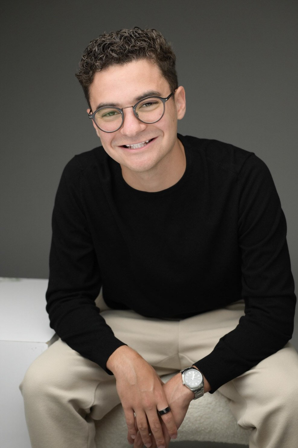 A young man with curly hair, glasses, and a big smile, wearing a black sweater, beige pants, and a watch, sitting against a gray background.