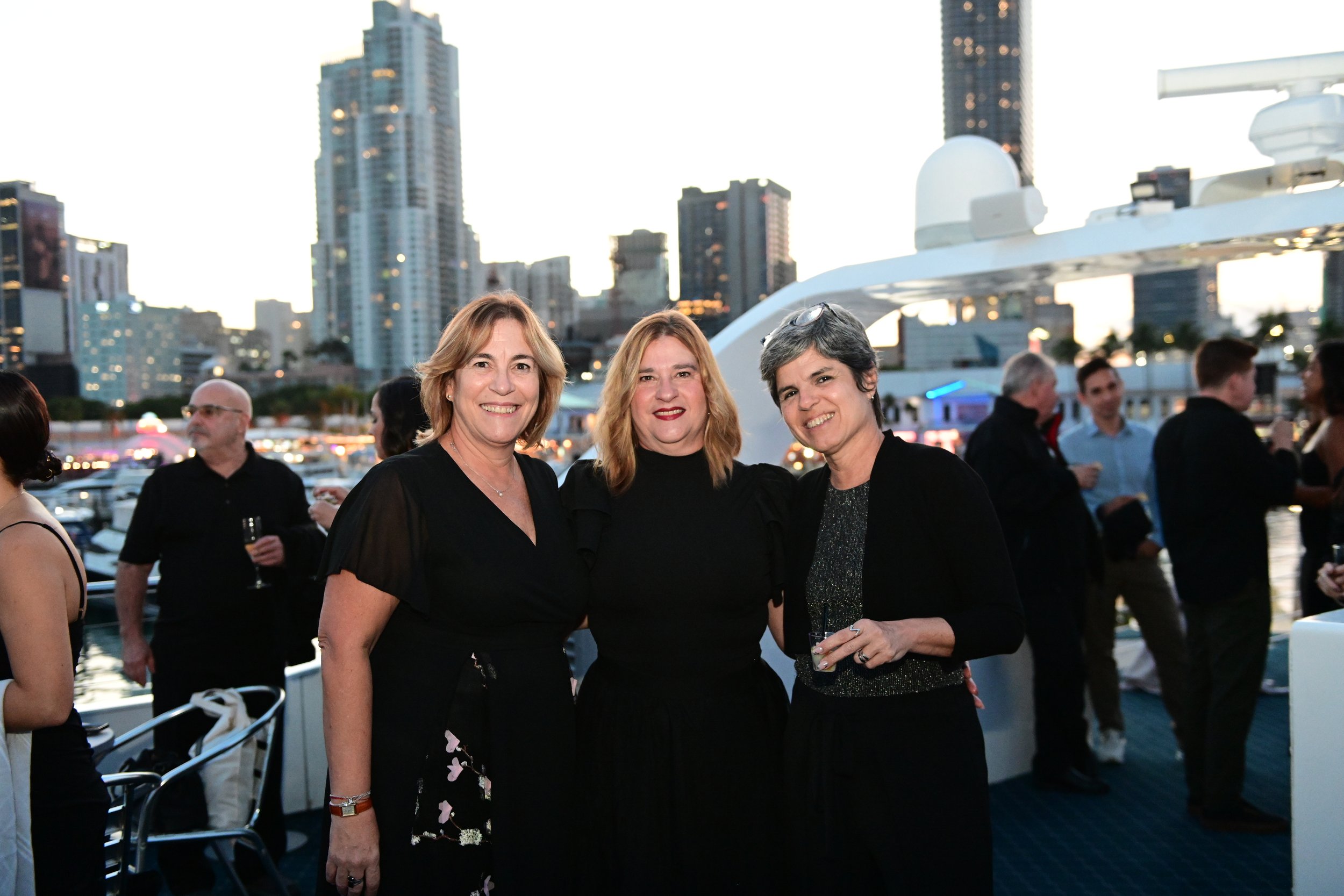 Three women standing together at an outdoor event with city skyscrapers and a marina in the background during sunset.