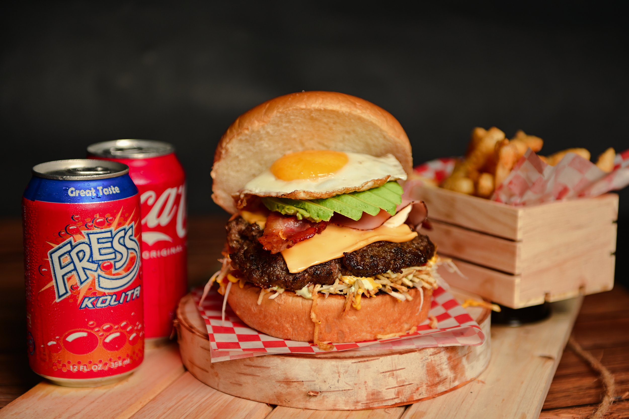 A large cheeseburger with lettuce, tomato, bacon, a fried egg, and multiple layers of meat and cheese on a bun, placed on a red-and-white checkered paper in a wooden basket. Two cans of soda, one blue and one red, are beside it, and a wooden basket o