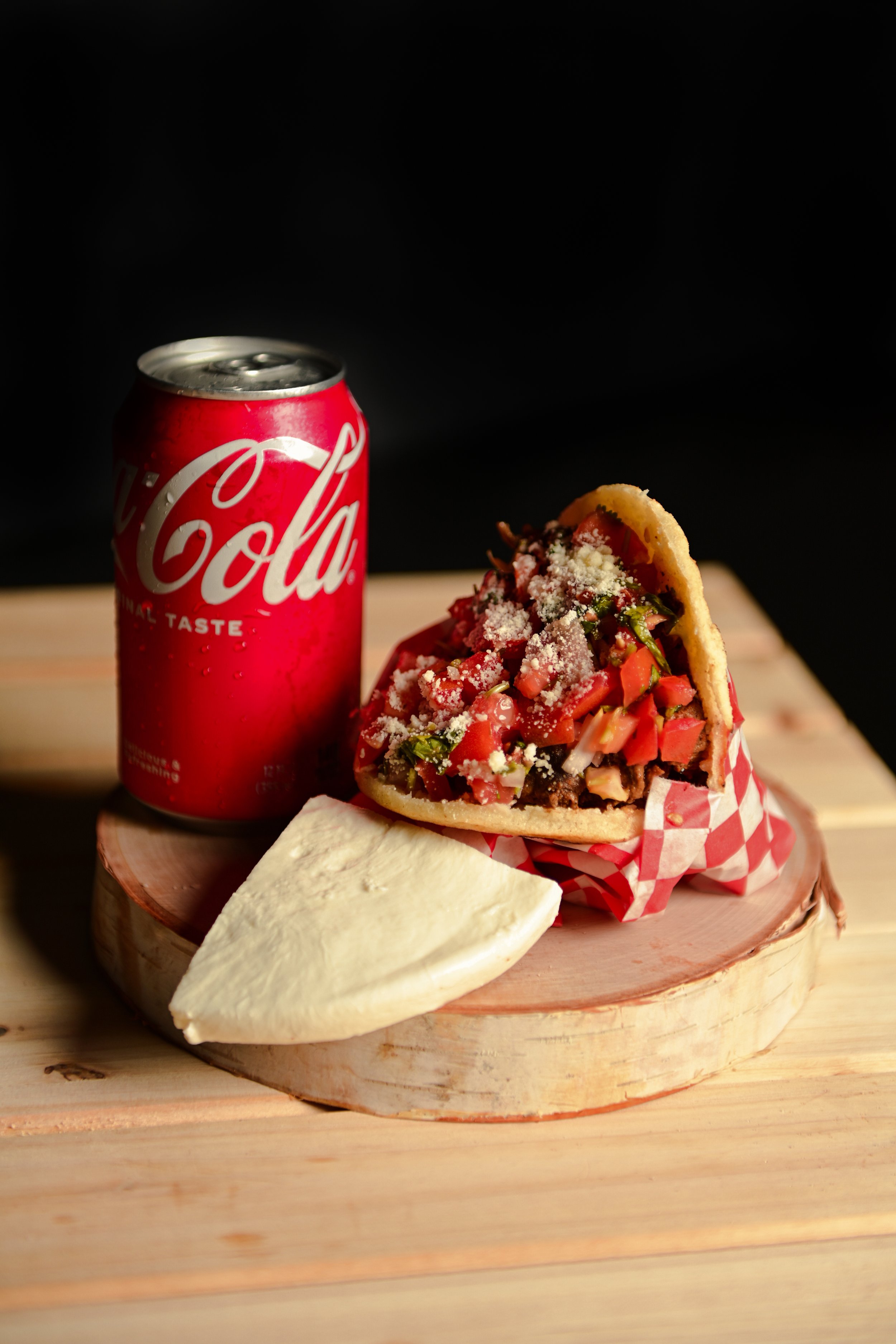 A red Coca-Cola can next to a chopped tomato salad in a taco, served on a wooden slice with a piece of white cheese on a wooden surface.