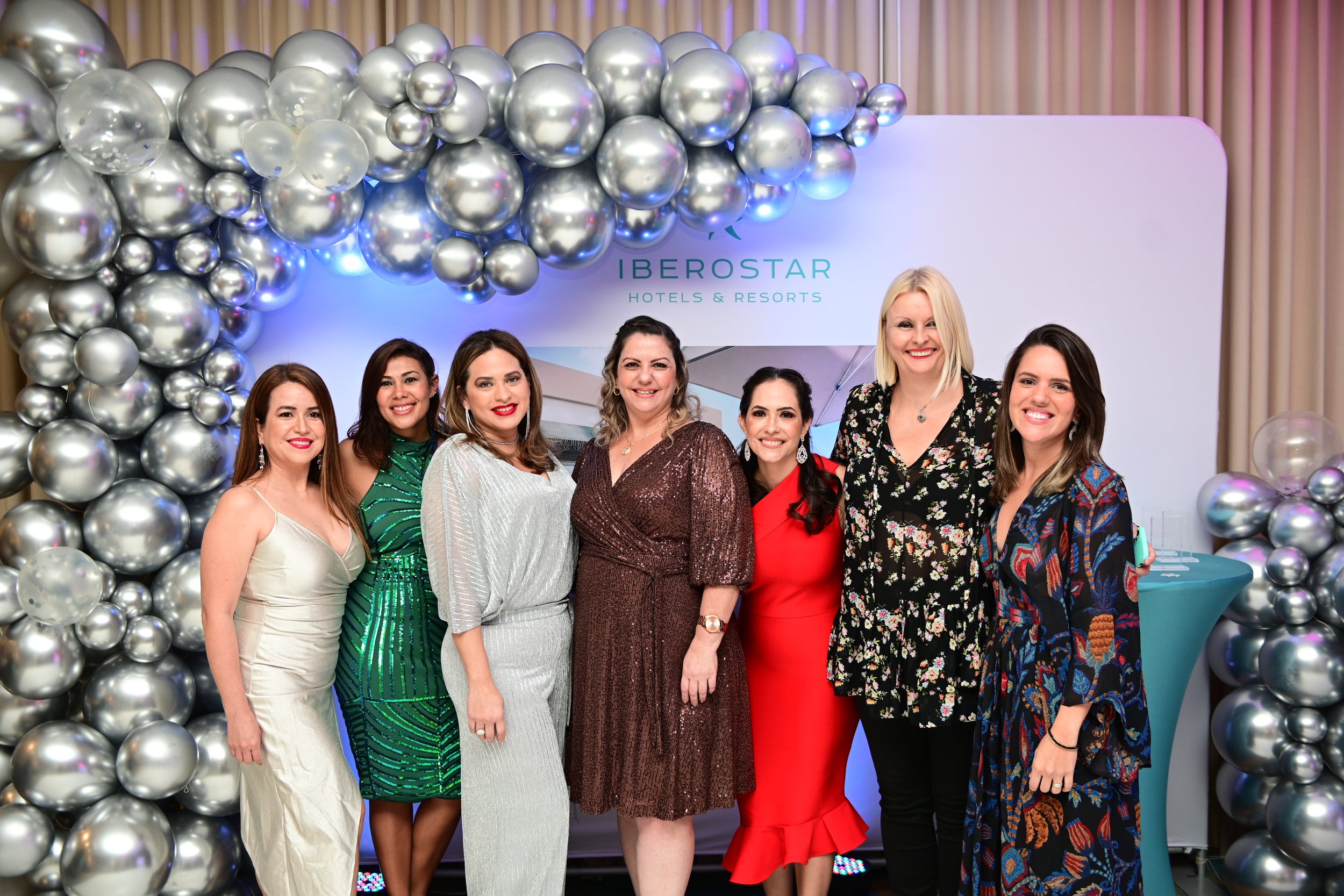 A group of six women in formal attire posing in front of a balloon decoration wall and a sign that reads 'Iberostar Hotels & Resorts'.
