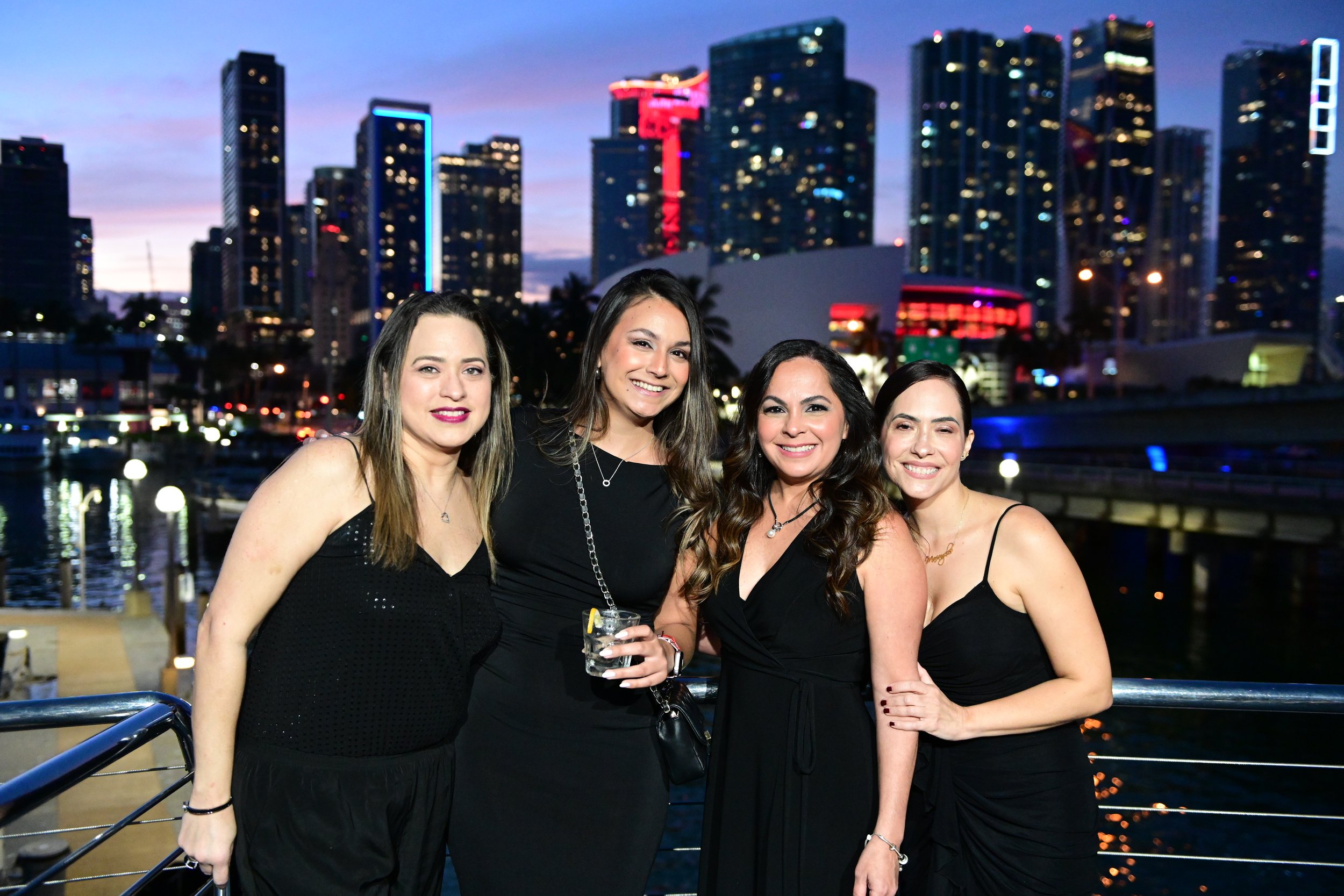 A group of four women in black dresses standing on a yacht at dusk, with a city skyline illuminated by city lights in the background.