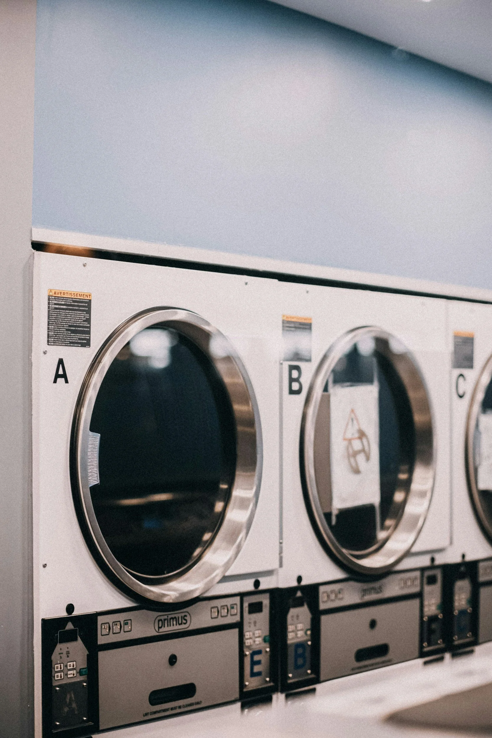A row of three white commercial laundry dryers with circular glass doors in a laundry room.