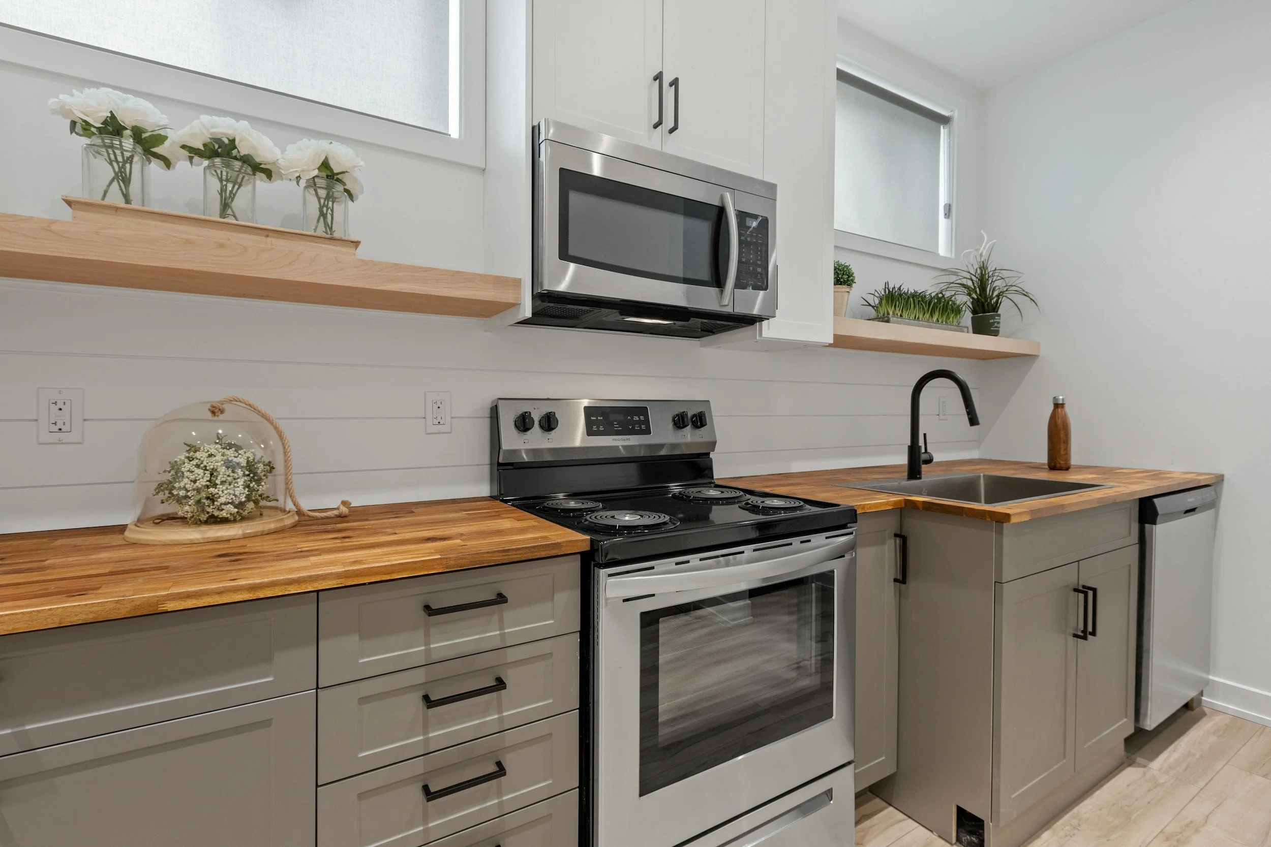 Modern kitchen with wood countertops, white cabinetry, stainless steel microwave and stove, black faucet, and green plants on open wooden shelves.