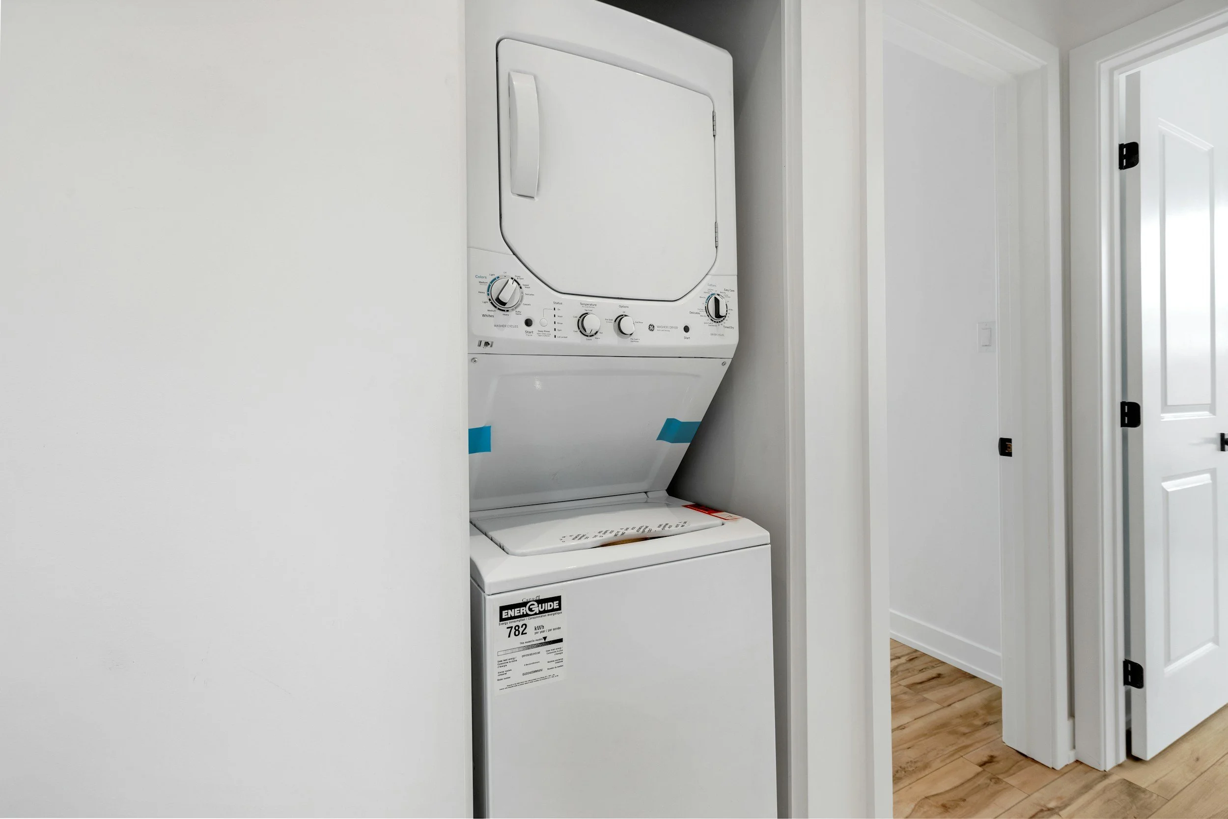 White stacked washer and dryer unit in small laundry closet with white walls and wooden floor