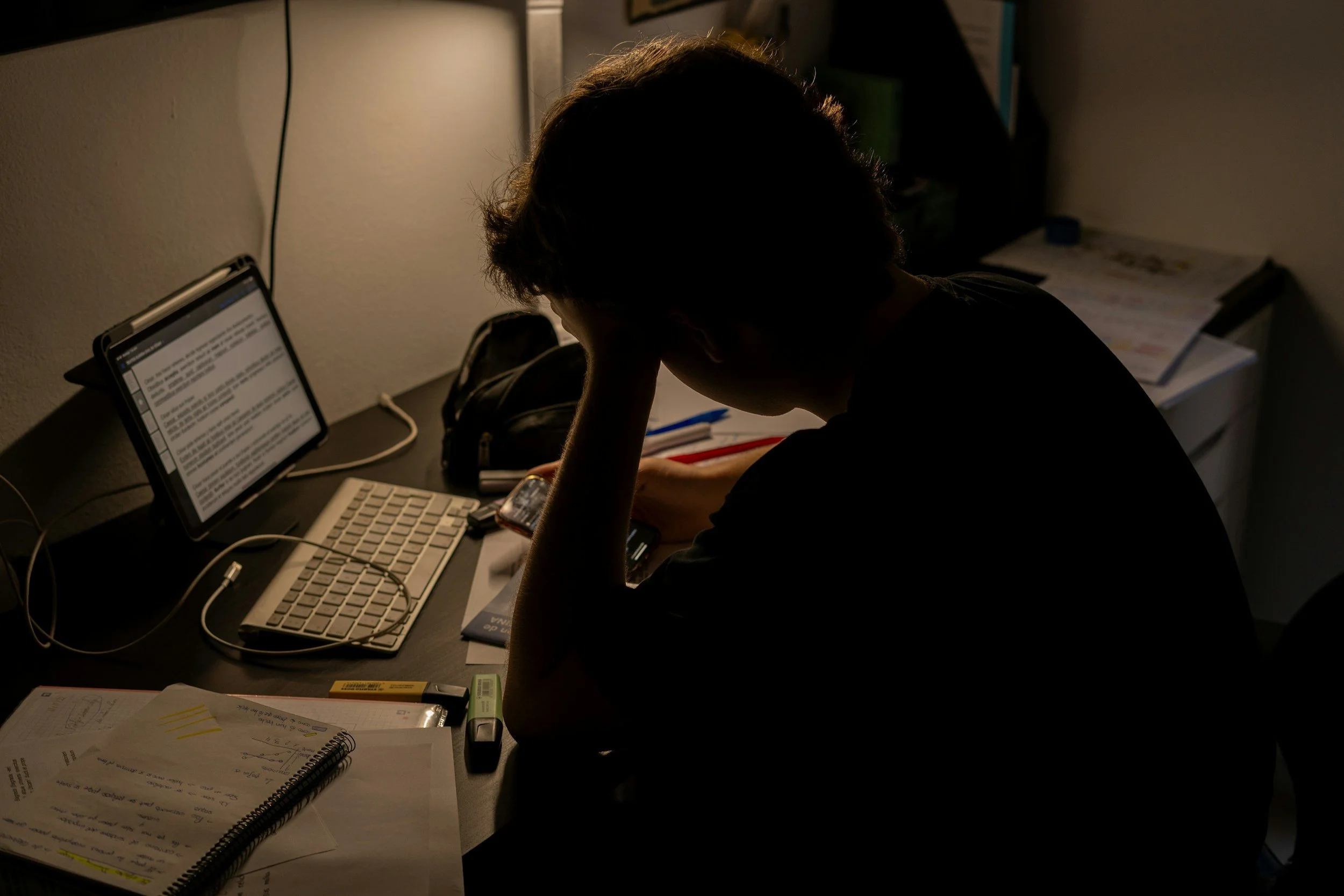 Person sitting at a cluttered desk, head resting in hand, illuminated by a computer screen in a dimly lit room.