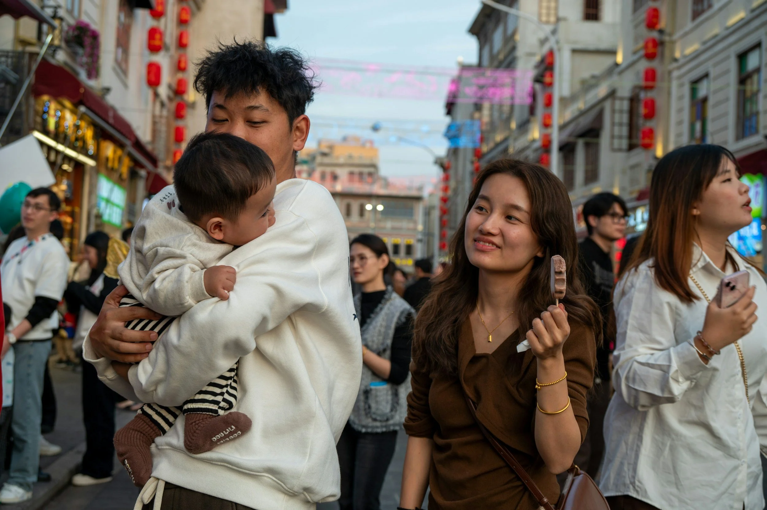 People on a busy street, some are holding hands, smiling, and talking, wearing casual jackets, with colorful buildings and lanterns in the background.