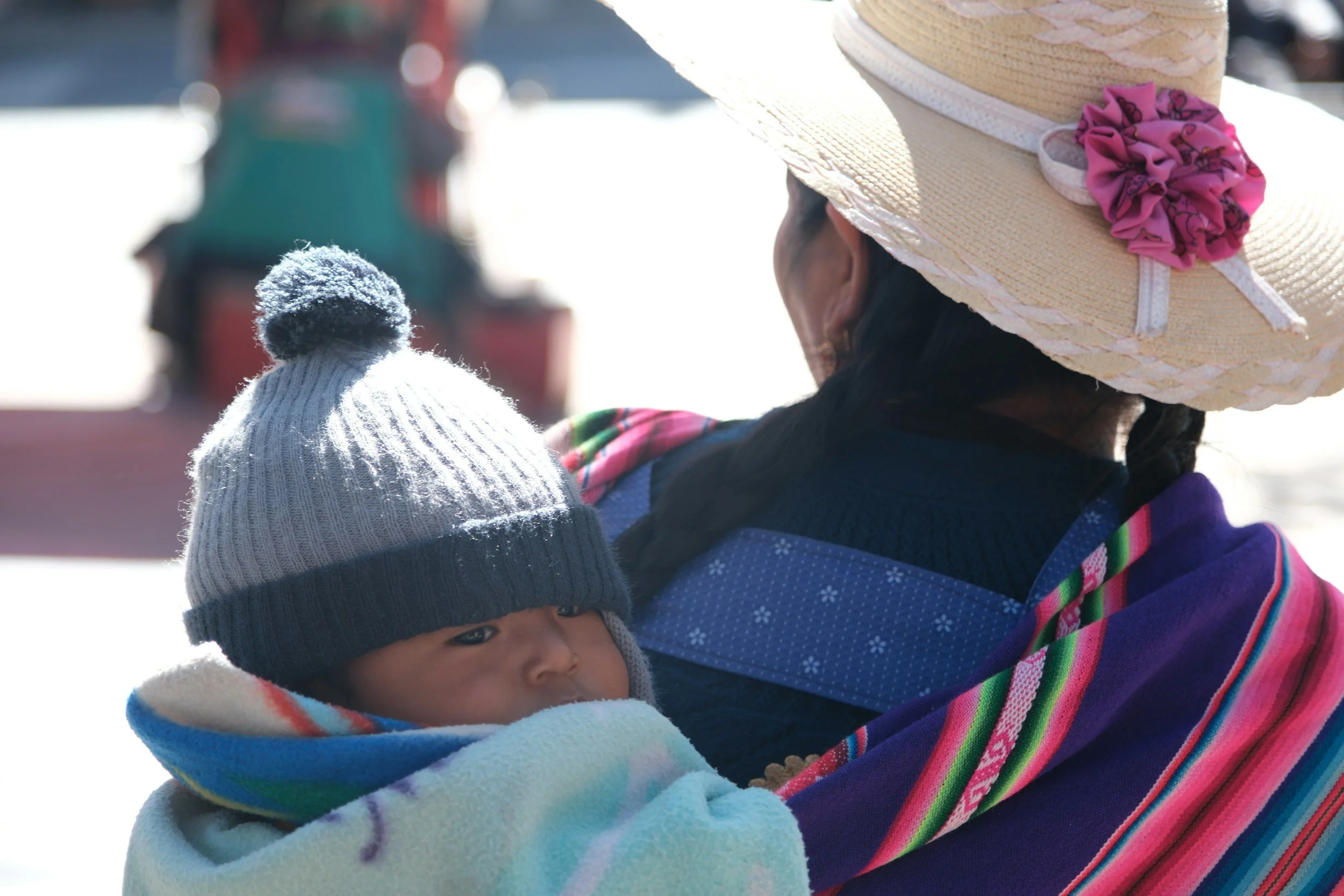 A woman holding a young child in cold weather. The woman wears a large sun hat with a pink flower decoration. The child is wrapped in a colorful striped blanket and wearing a gray knit hat with a pom-pom. Both are outdoors in the snow.