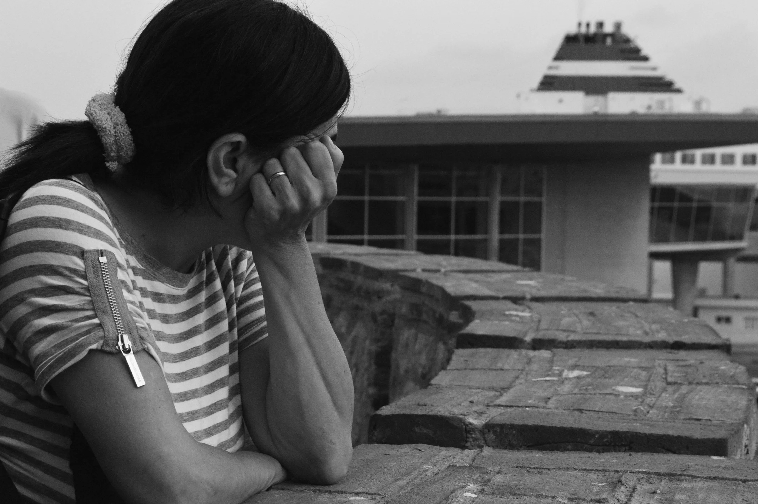 A woman with dark hair in a ponytail, wearing a striped shirt, sitting on a wooden bench with her face covered by her hands in front of a building with a pyramid-shaped roof.