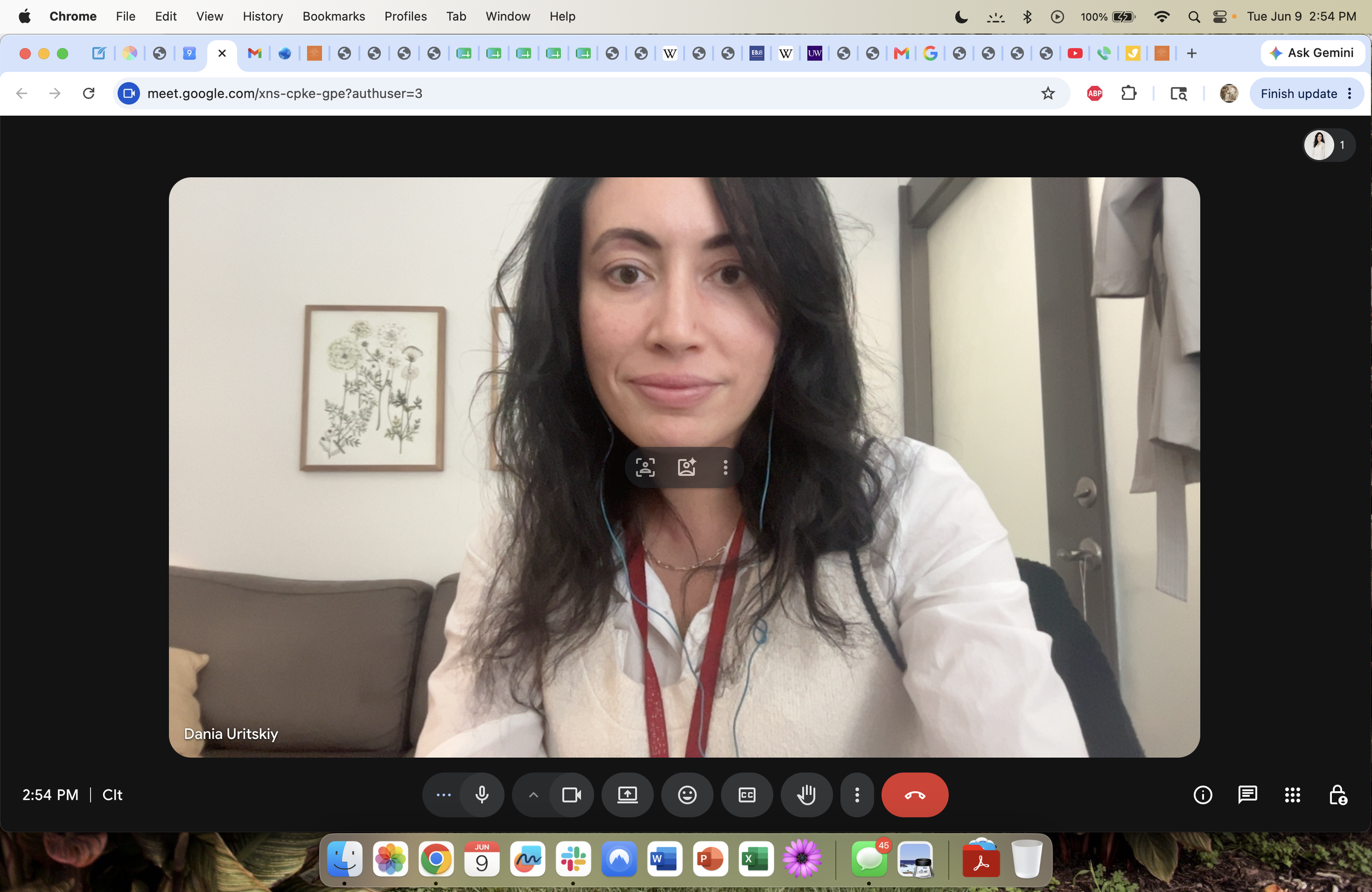 A woman with long dark curly hair and light skin, smiling slightly, in a video call. She is wearing a white shirt with a red lanyard and blue earbuds. The background shows a wall with a framed botanical print, part of a sofa with cushions, and a door with a coat hanging on it.