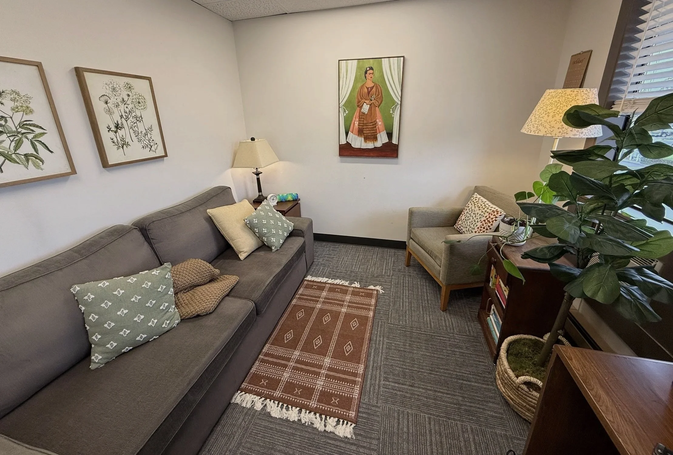 A cozy waiting room with a gray sofa, beige armchair, and wooden side table, decorated with framed botanical prints on the white walls, a rug, and potted plants by the window.