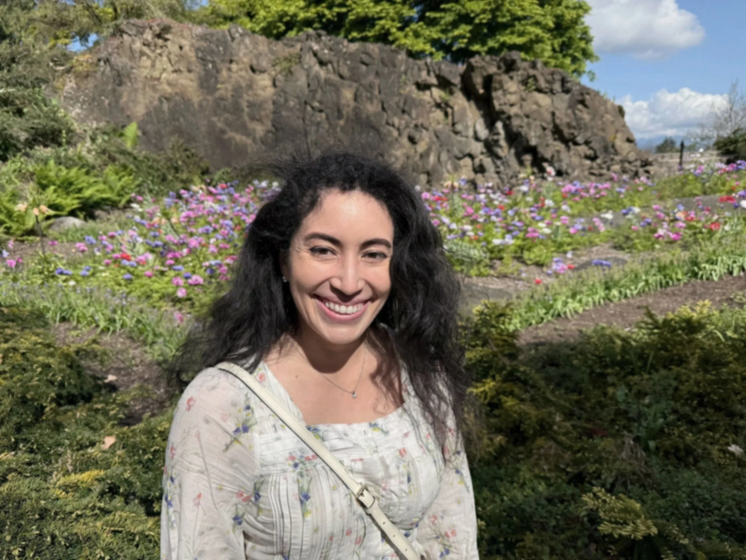 A woman with long dark hair smiling outdoors in front of a garden filled with pink, purple, and white flowers, with a rocky hill or cliff and green trees in the background.