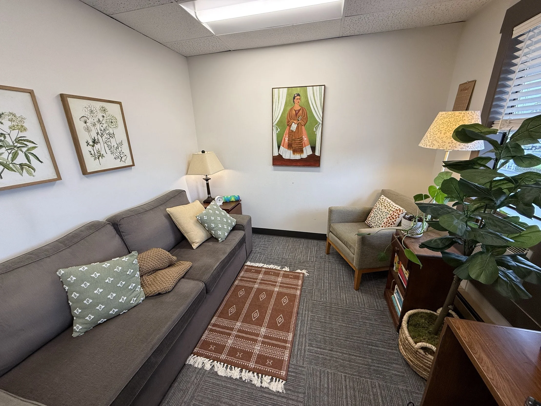 A cozy living room with a dark gray sofa decorated with patterned pillows, a small wooden side table with a lamp and clock, botanical artwork on the wall, a reddish-brown area rug, and a large potted plant near a window with blinds.