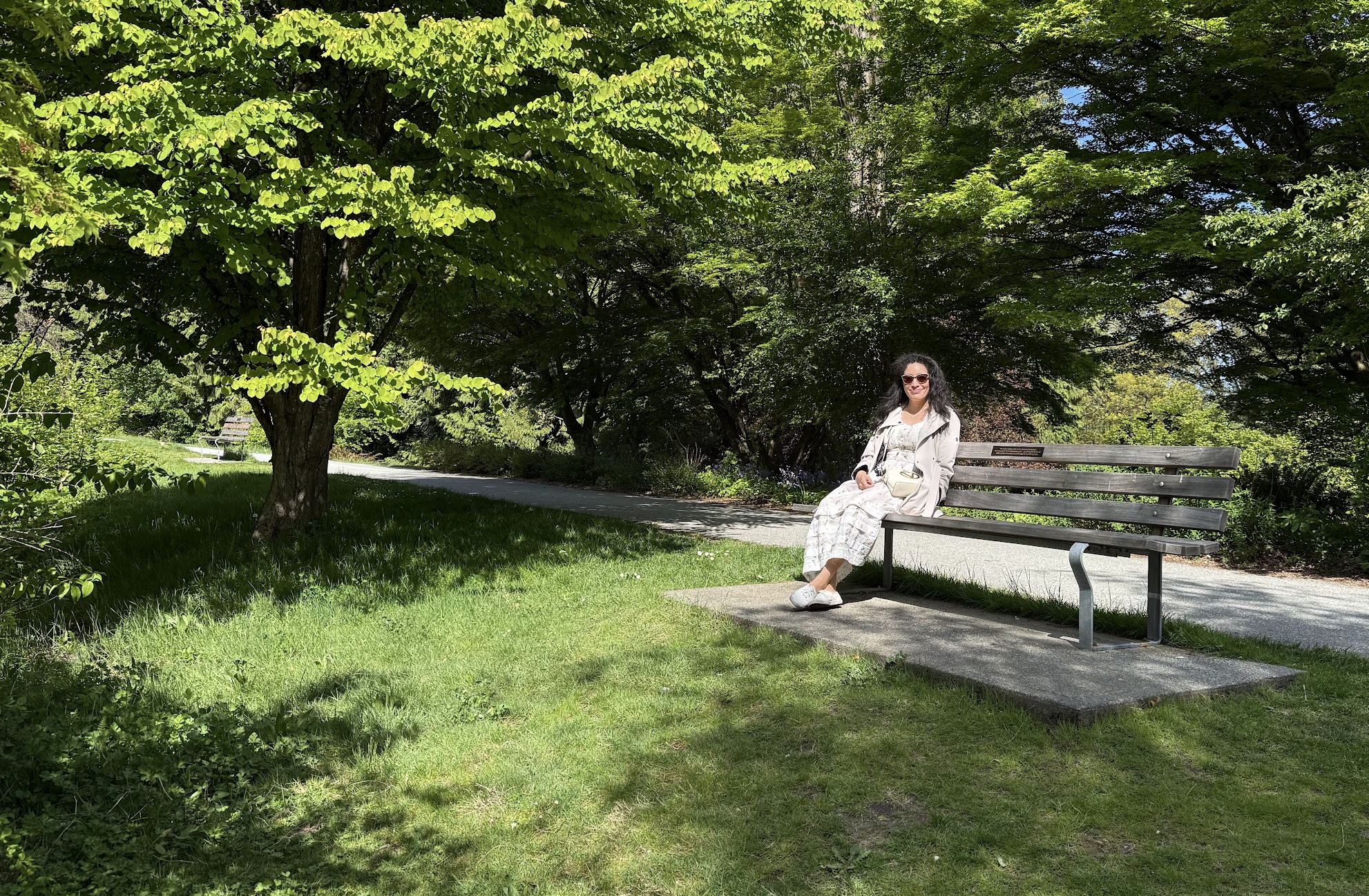 A woman with black curly hair wearing sunglasses, a light-colored dress, white sneakers, and a light-colored jacket is sitting on a park bench in a lush green park with trees and grass on a sunny day.