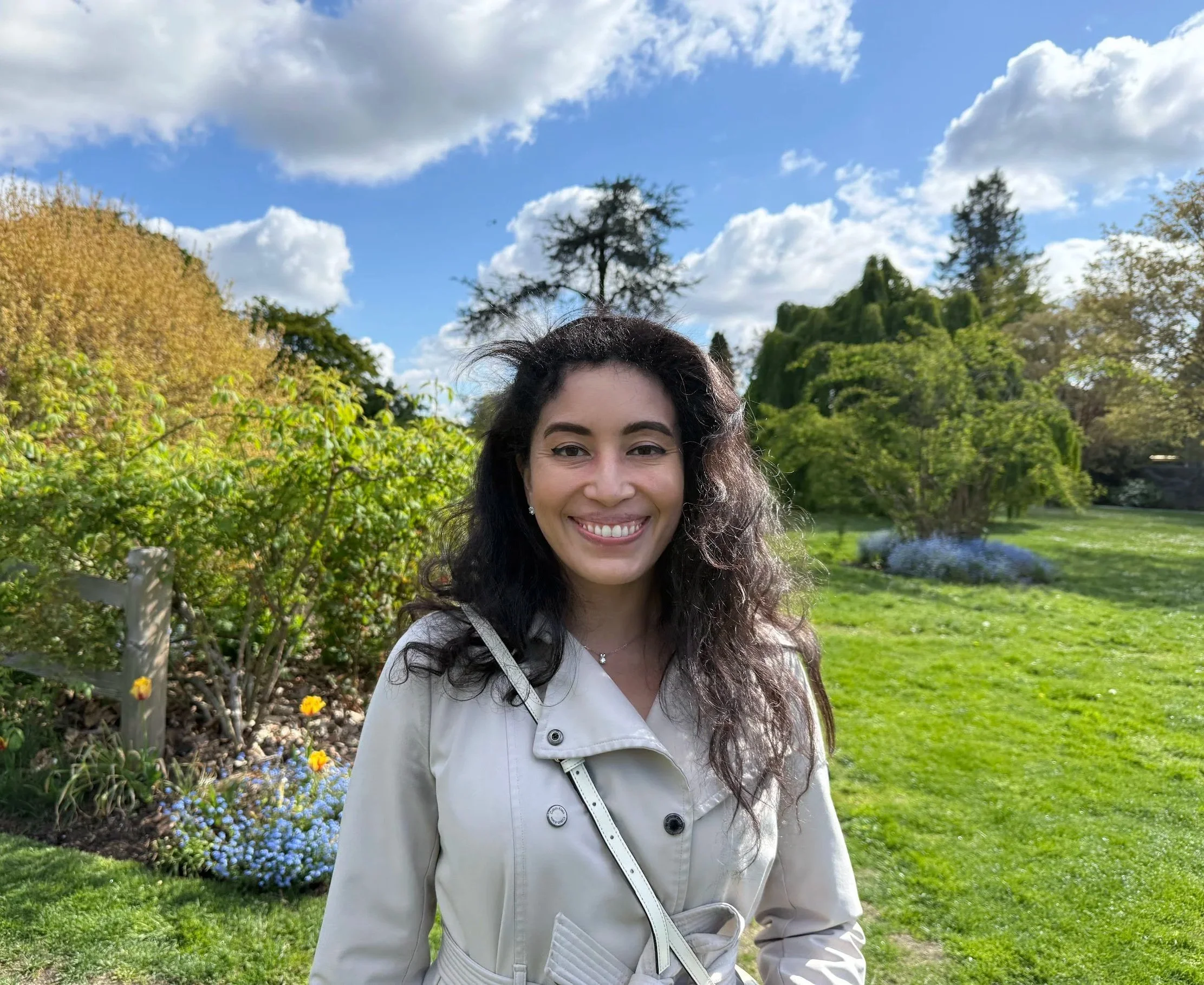 Smile of a woman with dark hair wearing a white jacket in a park with trees, flowers, and a partly cloudy sky.