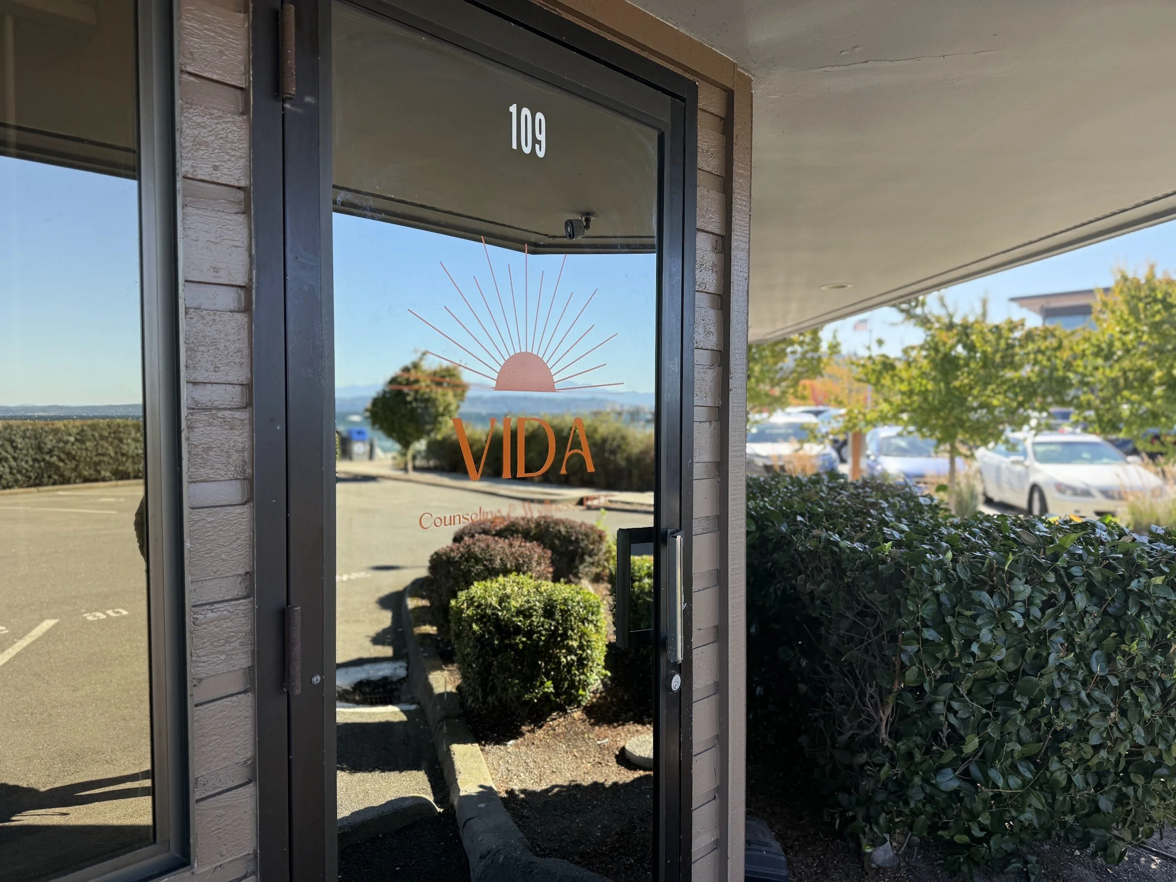 Storefront window with the number 109 and a sunburst logo, next to a landscaped parking lot and green bushes.