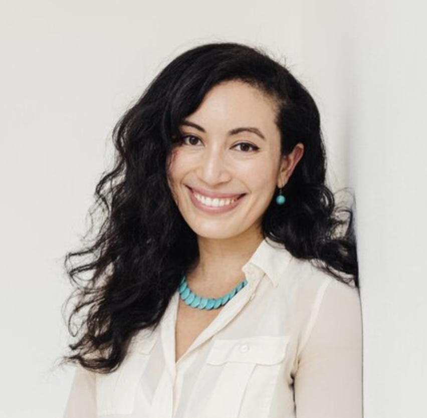 A smiling woman latina thrapist with dark curly hair. She is warm and welcoming. She is a professional therapist. The photo is taken in downtown Seattle WA in a therapy office.