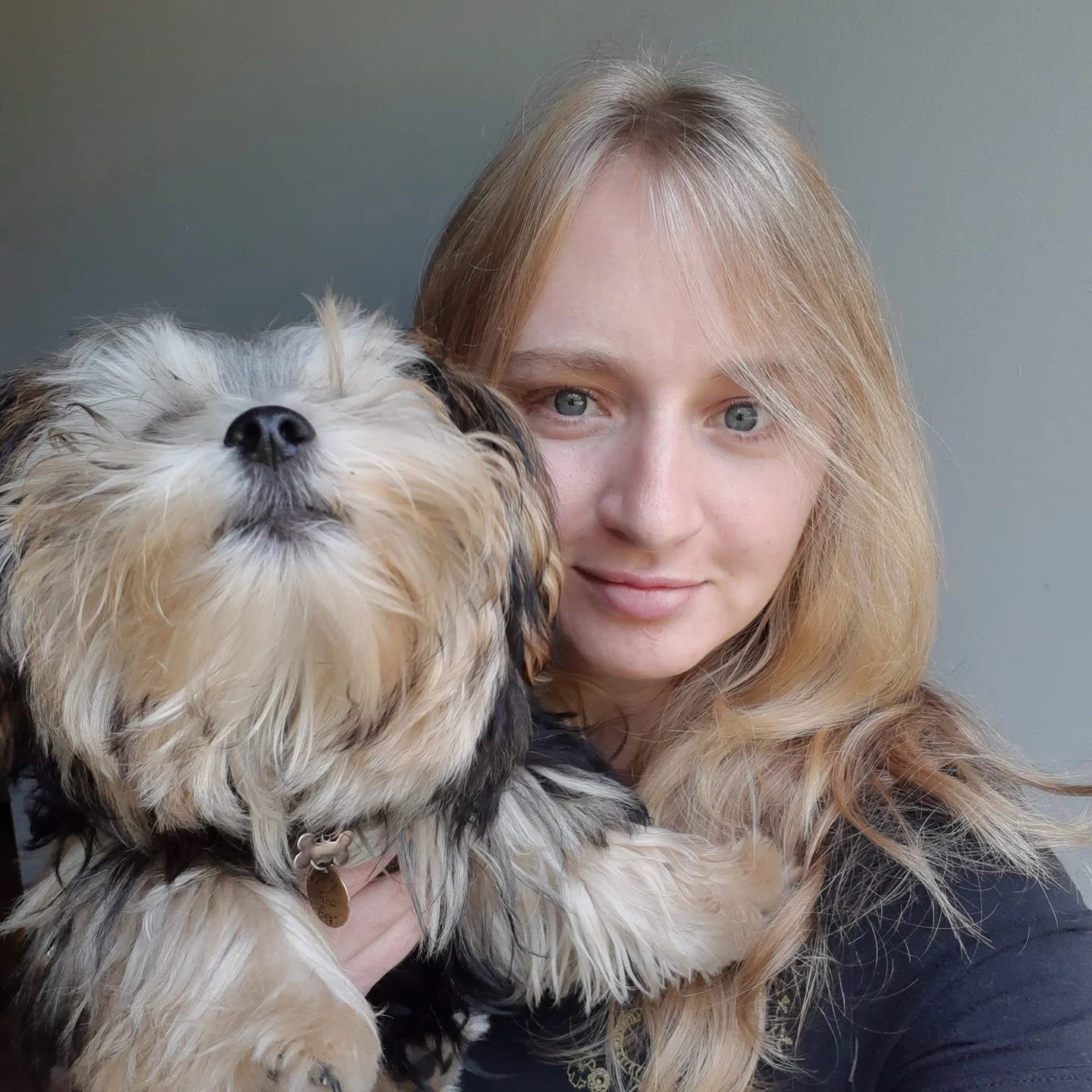A young woman with blonde hair and blue eyes smiling while holding a fluffy dog with a black and tan coat close to her face.