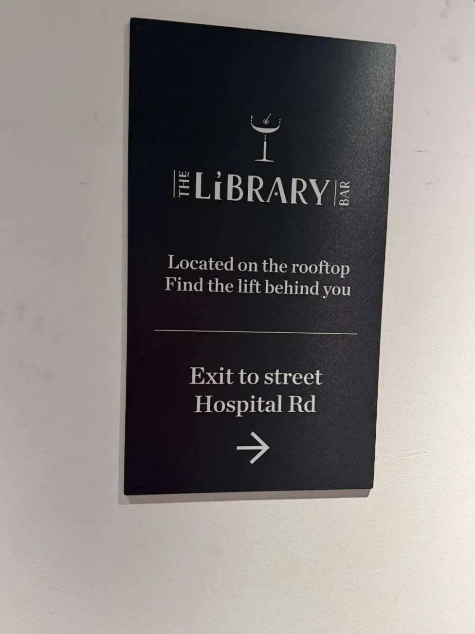 Sign for The Library Bar with directions, located on the rooftop, indicating to find the lift behind you and exit to street on Hospital Road, with an arrow pointing to the right.