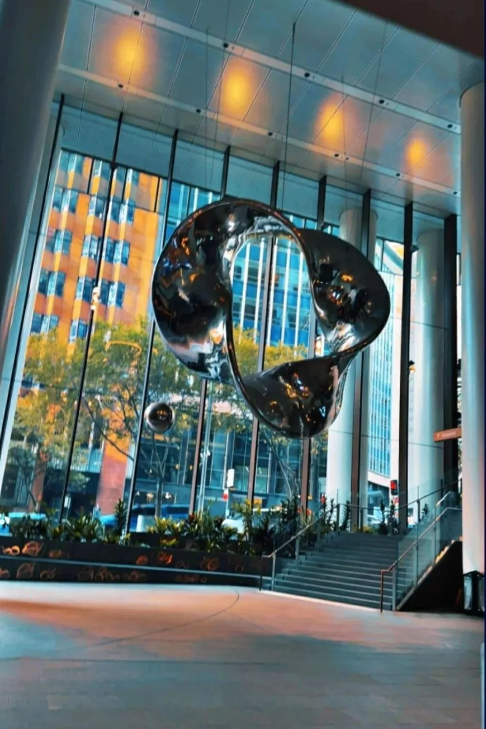 Modern lobby with large glass windows, a metallic abstract sculpture hanging from the ceiling, trees and skyscrapers outside, and a staircase with plants near the entrance.