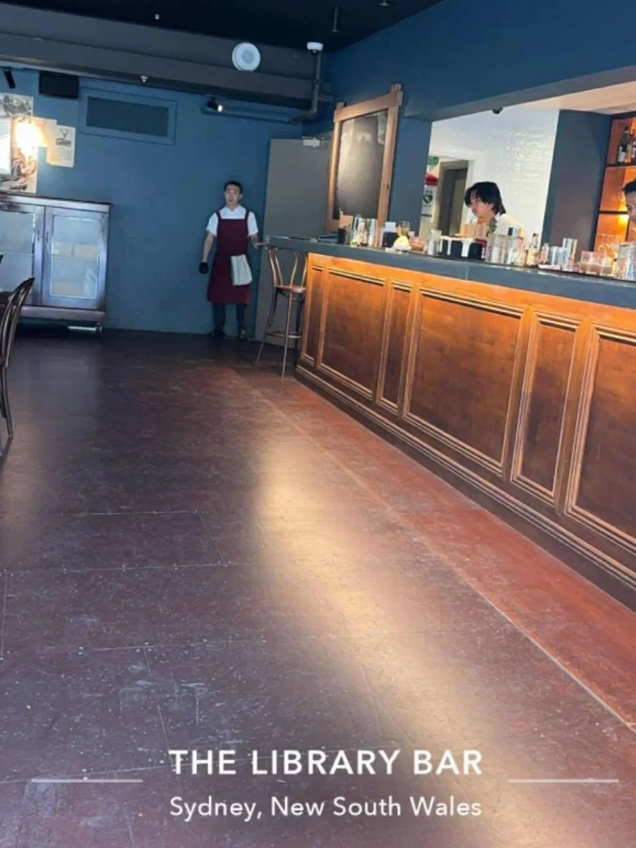 Empty bar area at The Library Bar in Sydney, New South Wales, with a bartender behind the counter and a staff member standing near the wall. The bar has wooden paneling and various bottles and glasses on the counter.