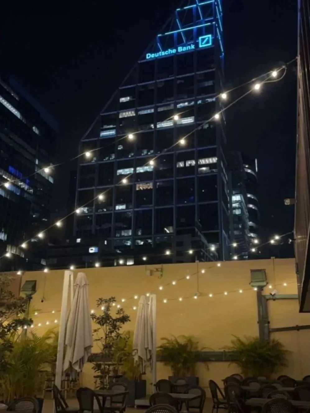 Nighttime view of a city rooftop with string lights, outdoor seating, and a high-rise building with neon signs, including a Deutsche Bank logo.