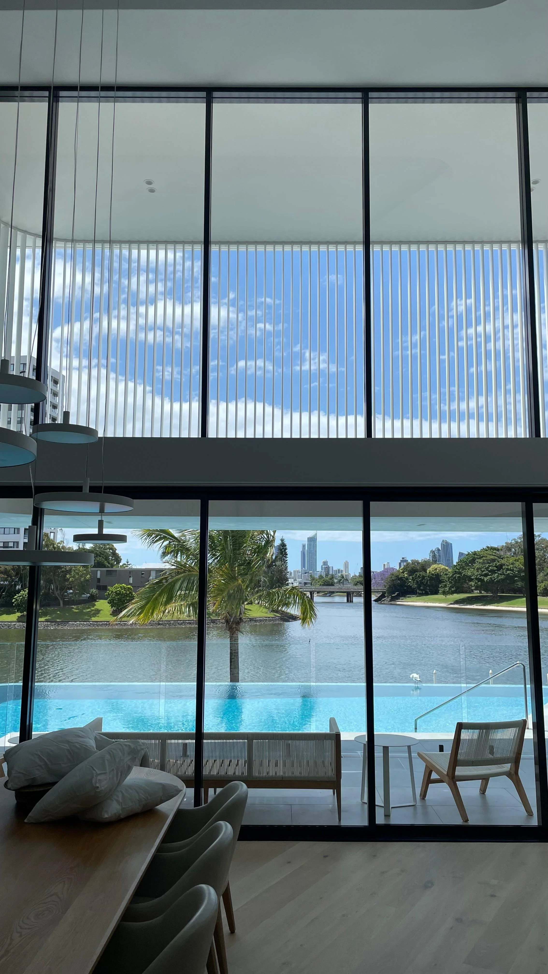 Modern living room with floor-to-ceiling windows overlooking a river, city skyline, pool, and outdoor seating area with a palm tree.