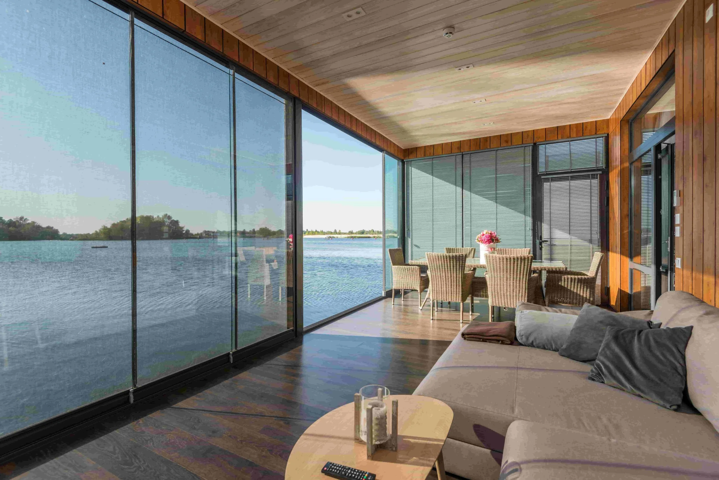 A cozy living room with floor-to-ceiling windows overlooking a lake, with a beige sofa, a small wooden table with candles and a TV remote, and a dining area with a round table and wicker chairs, decorated with a pink flower vase.