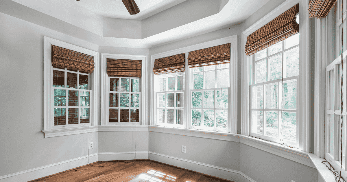A bright, empty room with large windows and wooden blinds, white walls, and hardwood flooring.