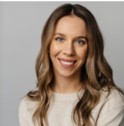 A young woman with long wavy brown hair smiling at the camera, wearing a light-colored top against a gray background.