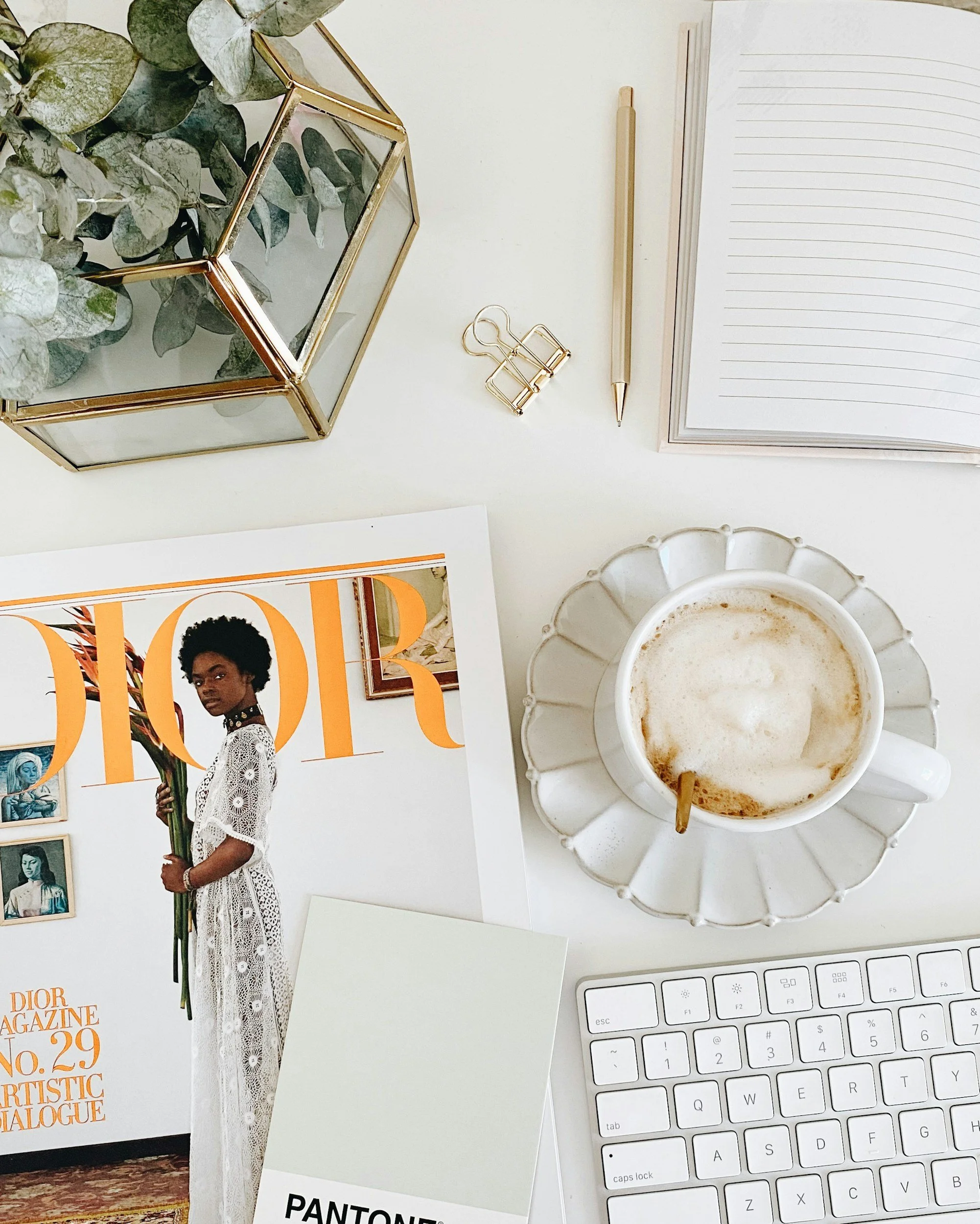 A flat lay of a workspace including a Dior magazine, a Pantone color swatch, a white keyboard, a cup of coffee with foam in a clear glass saucer, an open notebook, gold binder clips, a gold pen, and a geometric glass terrarium with green eucalyptus leaves.