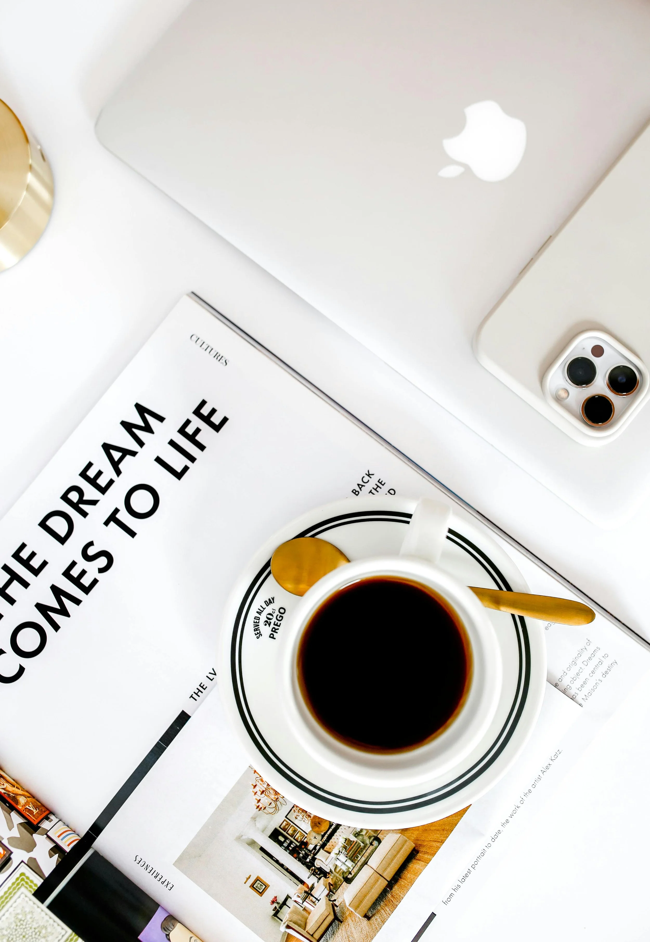 Top view of a workspace with a cup of coffee, a closed silver MacBook, a white smartphone with three camera lenses, a magazine, and a gold-colored object, all arranged on a white surface.