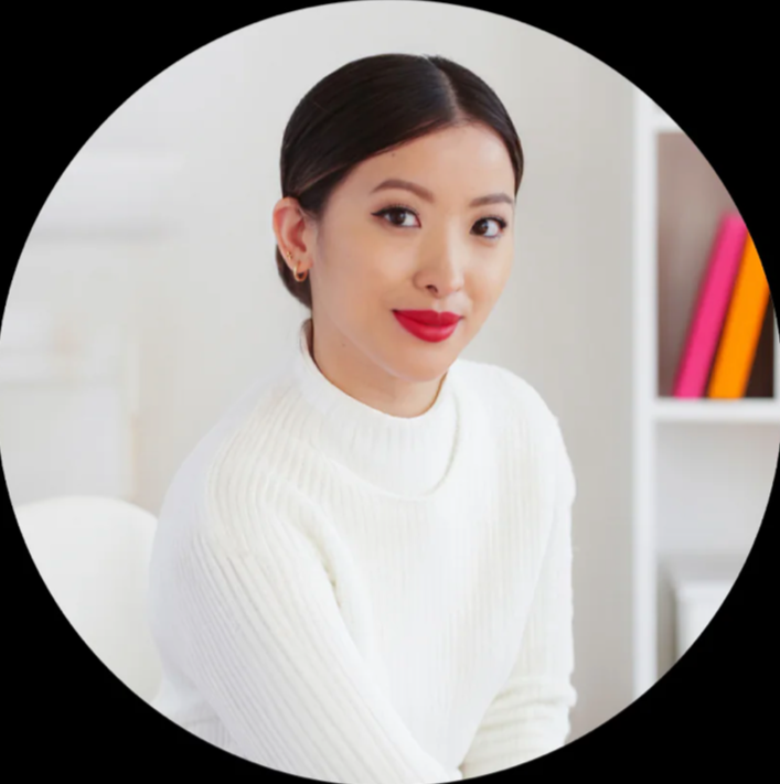 A woman with dark hair, wearing a white sweater and red lipstick, sitting in a bright room with a white bookshelf and colorful books in the background.