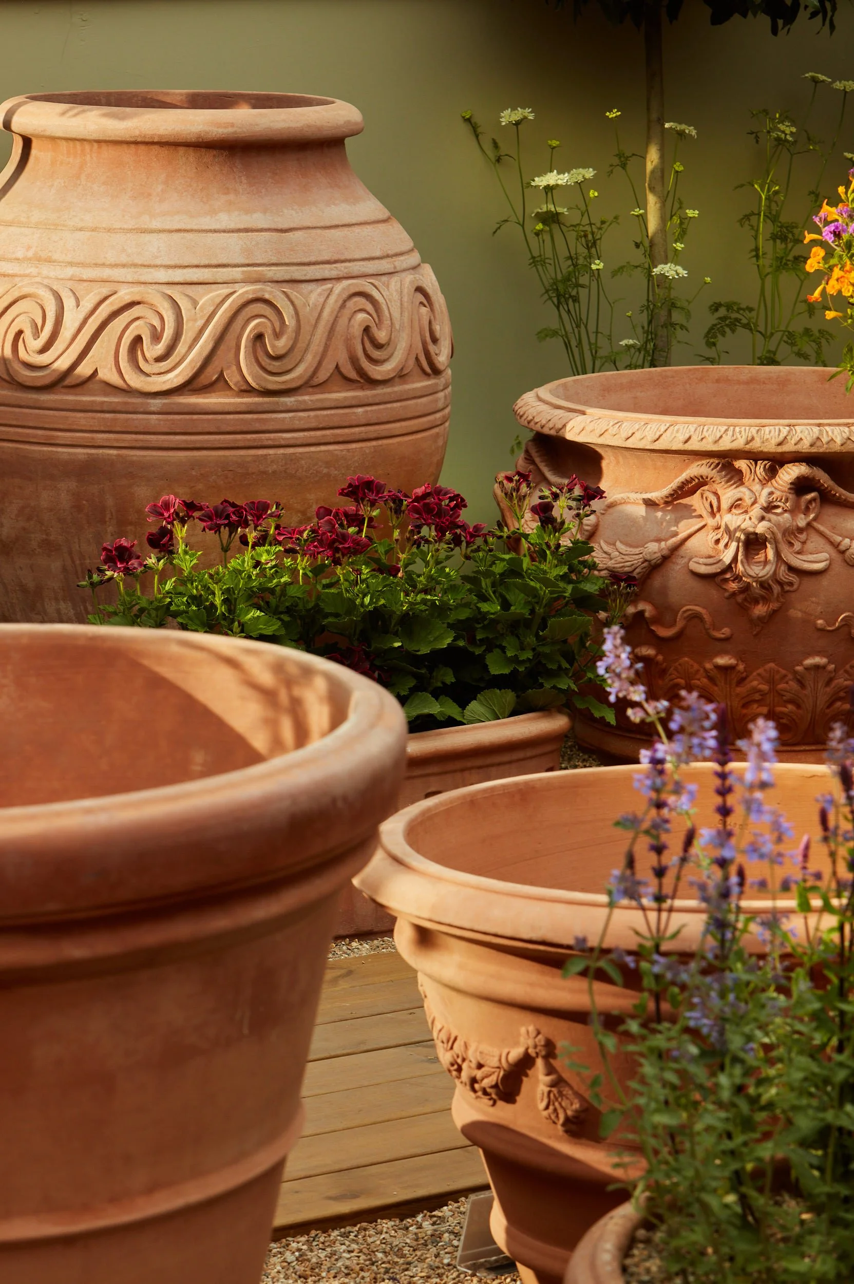 Multiple large decorative terracotta garden planters with various engraved and ornate designs, some filled with colorful flowering plants, on a wooden deck.