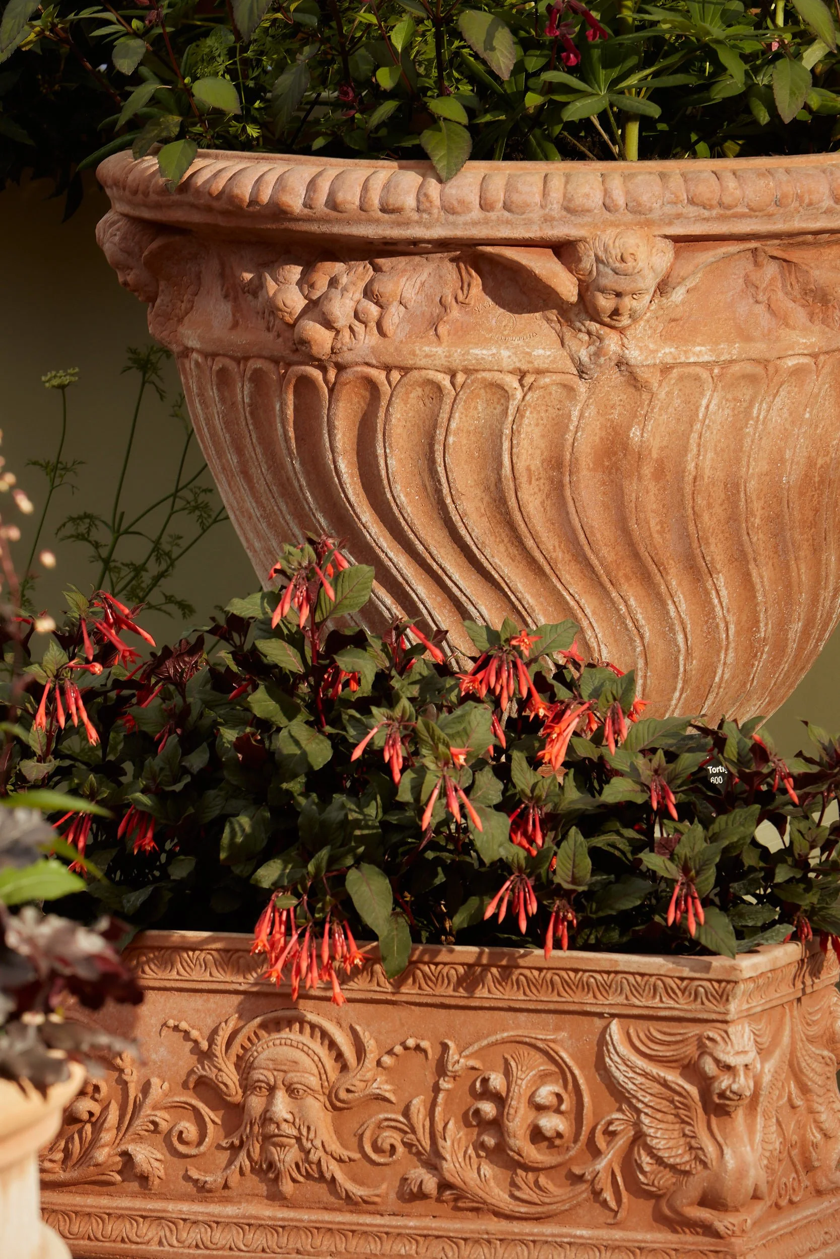 Decorative terracotta planters with intricate carvings, containing lush green and red flowering plants.