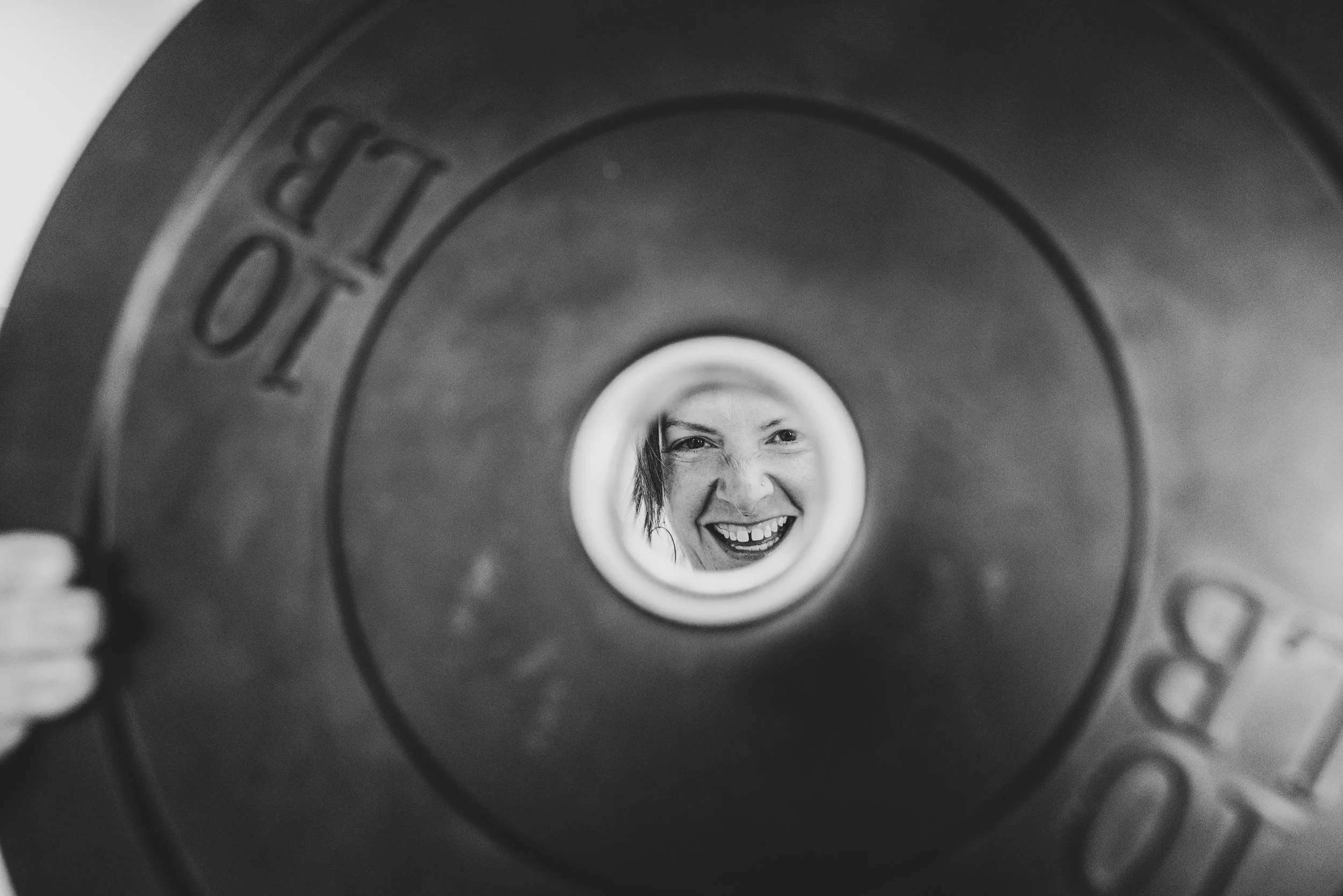 A smiling woman with short hair seen through the center hole of a weight plate.
