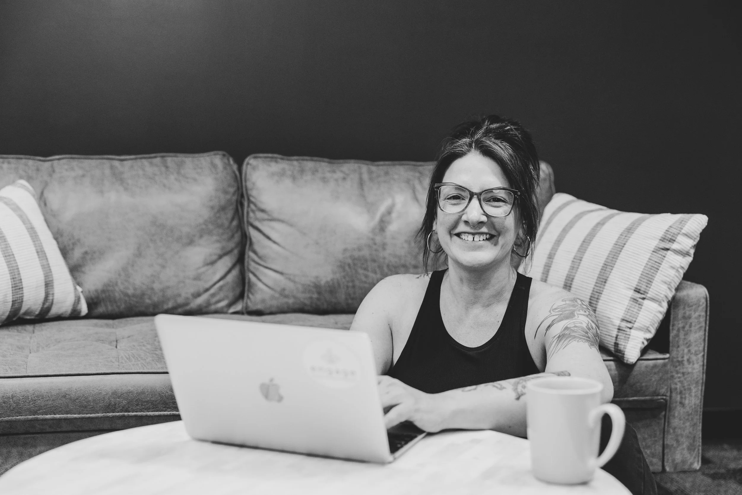 A woman with glasses and tattoos sitting in front of a laptop on a coffee table, smiling at the camera, with a mug beside her on the table and a sofa with striped pillows in the background.
