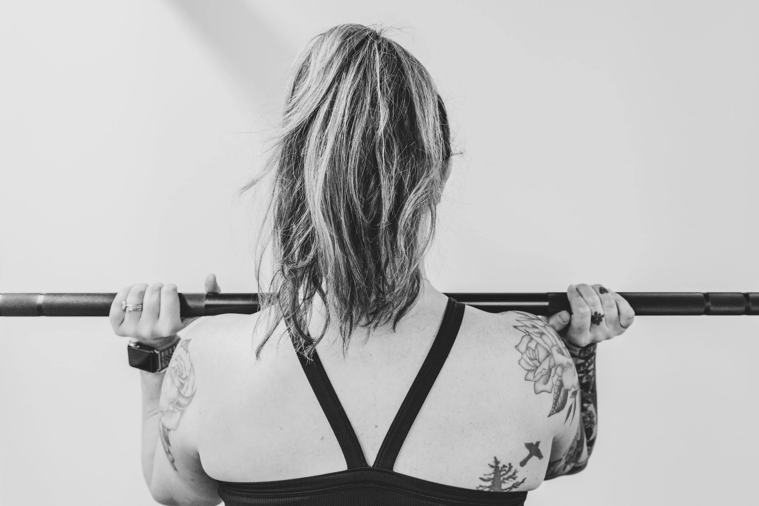 Back view of a woman with wavy hair holding a barbell across her shoulders, tattoos on her arms and shoulder, wearing a sports bra.