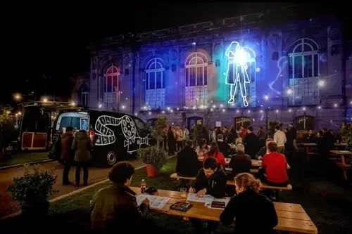 Une scène nocturne avec des personnes assises à des tables en extérieur, un bâtiment en arrière-plan avec des projections de lumières colorées et une grande figure humaine lumineuse.