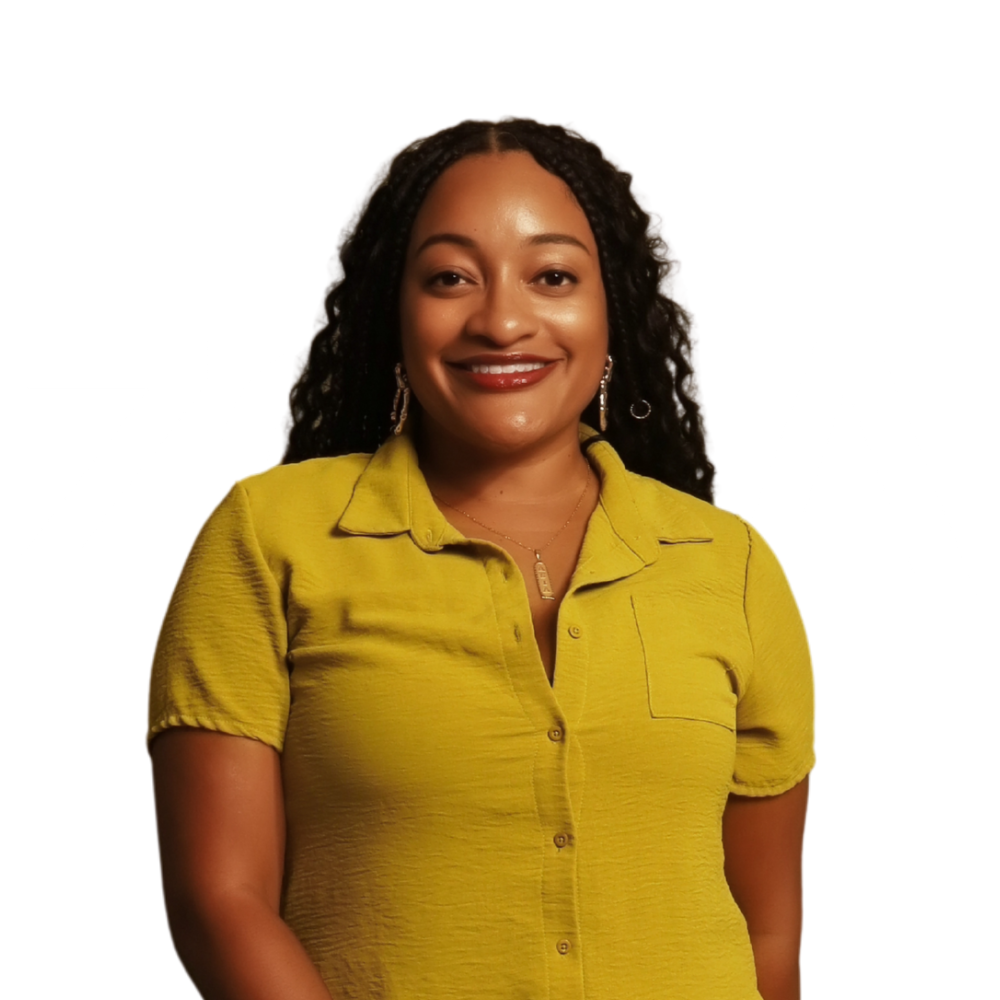 A young woman with braided hair, wearing a brown blazer over a white top, gold hoop earrings, a gold necklace with a rectangular pendant, smiling at the camera.