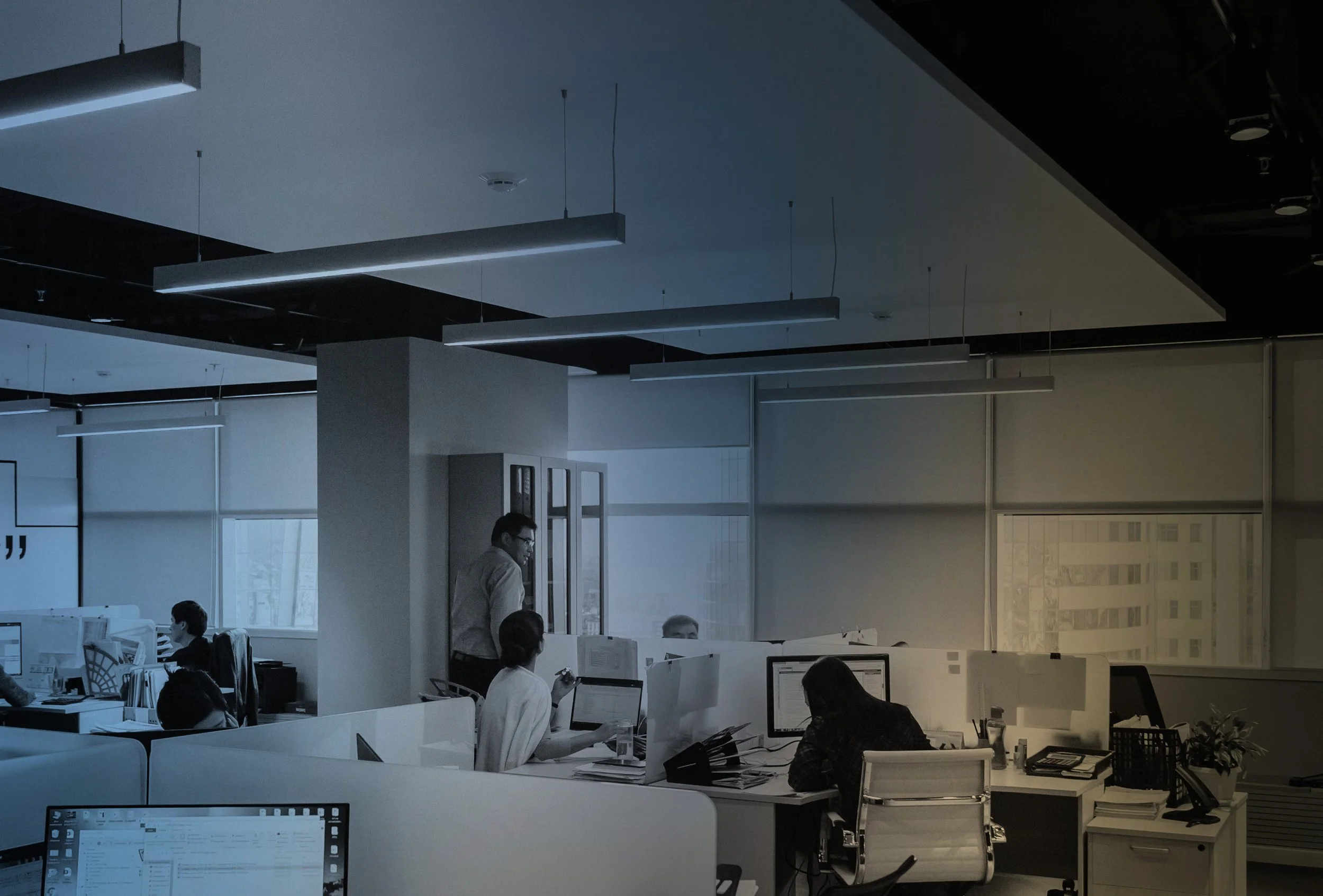 Bureaux modernes avec employés travaillant sur leurs ordinateurs, éclairage suspendu au plafond et fenêtres laissant entrer la lumière naturelle.