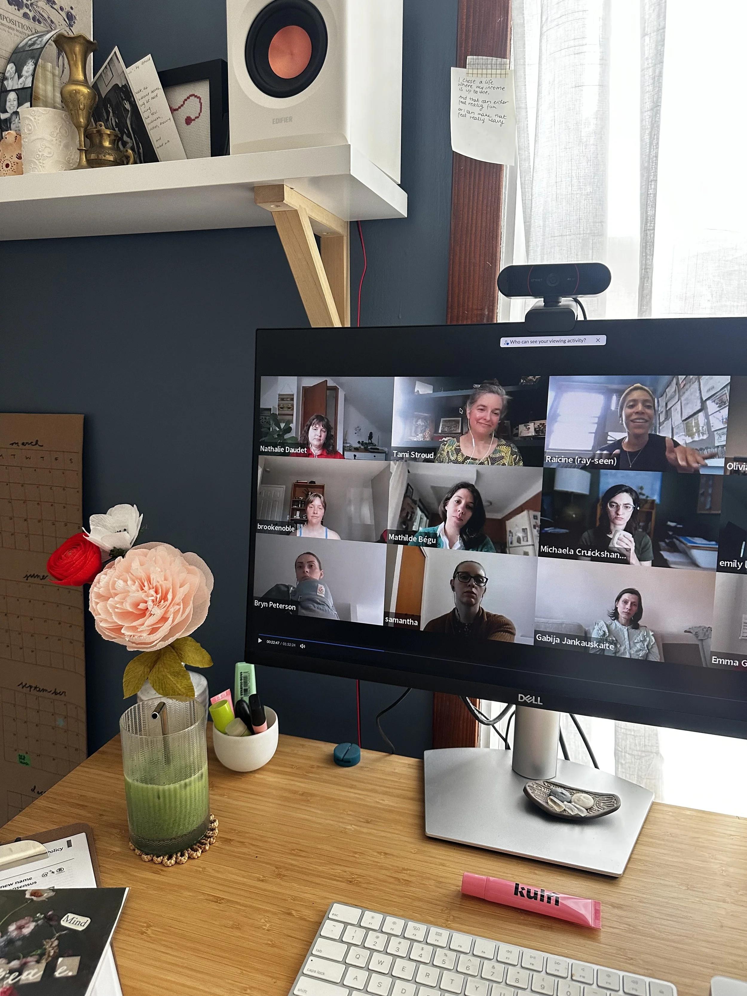 A workspace with a wooden desk holding a clear green glass vase with pastel-colored artificial flowers, a white keyboard, a pink lipstick, a white mouse pad, and decorative rocks. In the background, a computer monitor shows a video call with several participants. To the left of the desk, there's a calendar, and above, a shelf with framed photos, a gold vase, and a speaker. A white note with handwritten text is tacked to the wall near a window with sheer curtains.